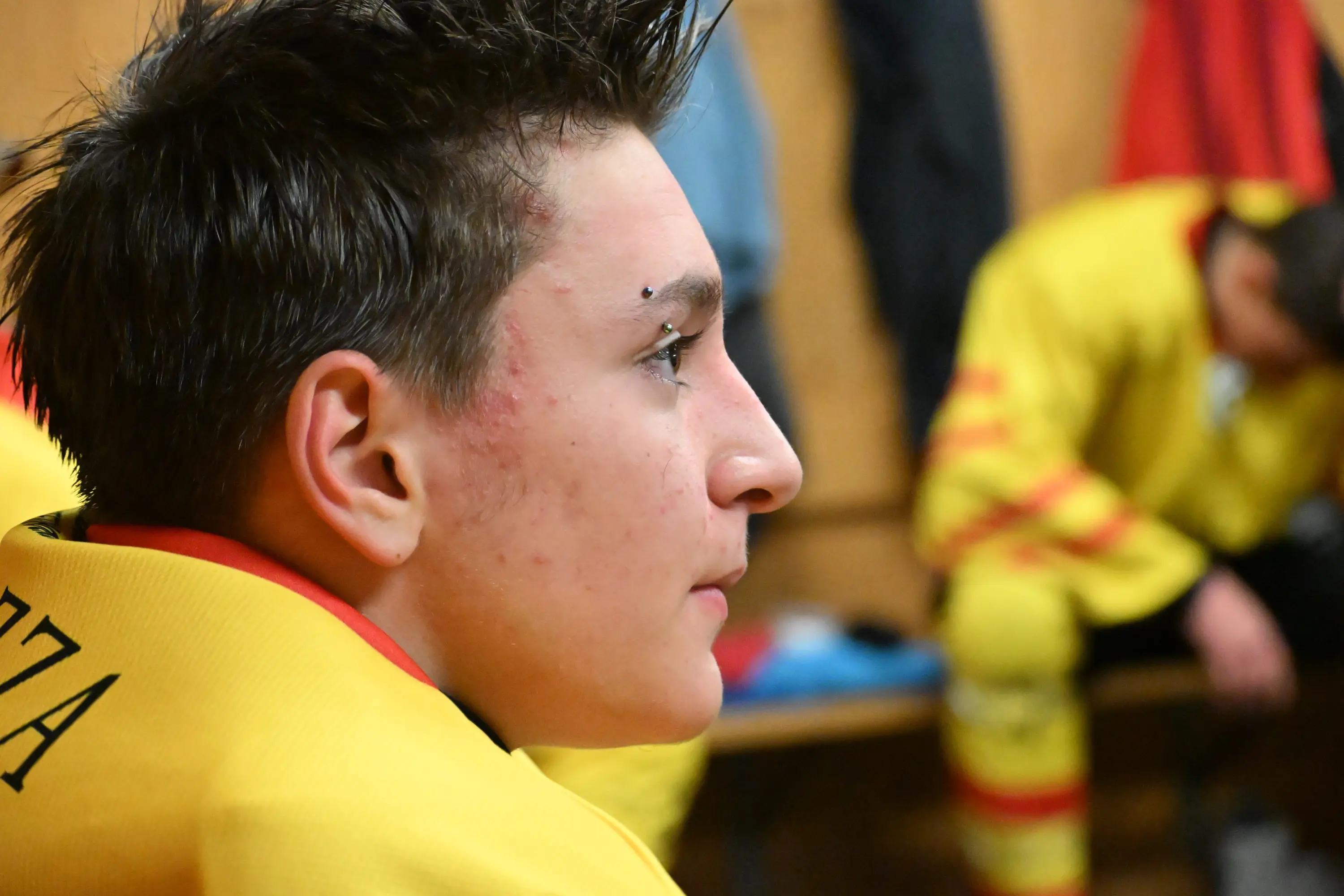 Ritratto di un giovane atleta con capelli in stile mohawk e un piercing, in attesa in uno spogliatoio. Indossa una maglietta gialla e sembra concentrato