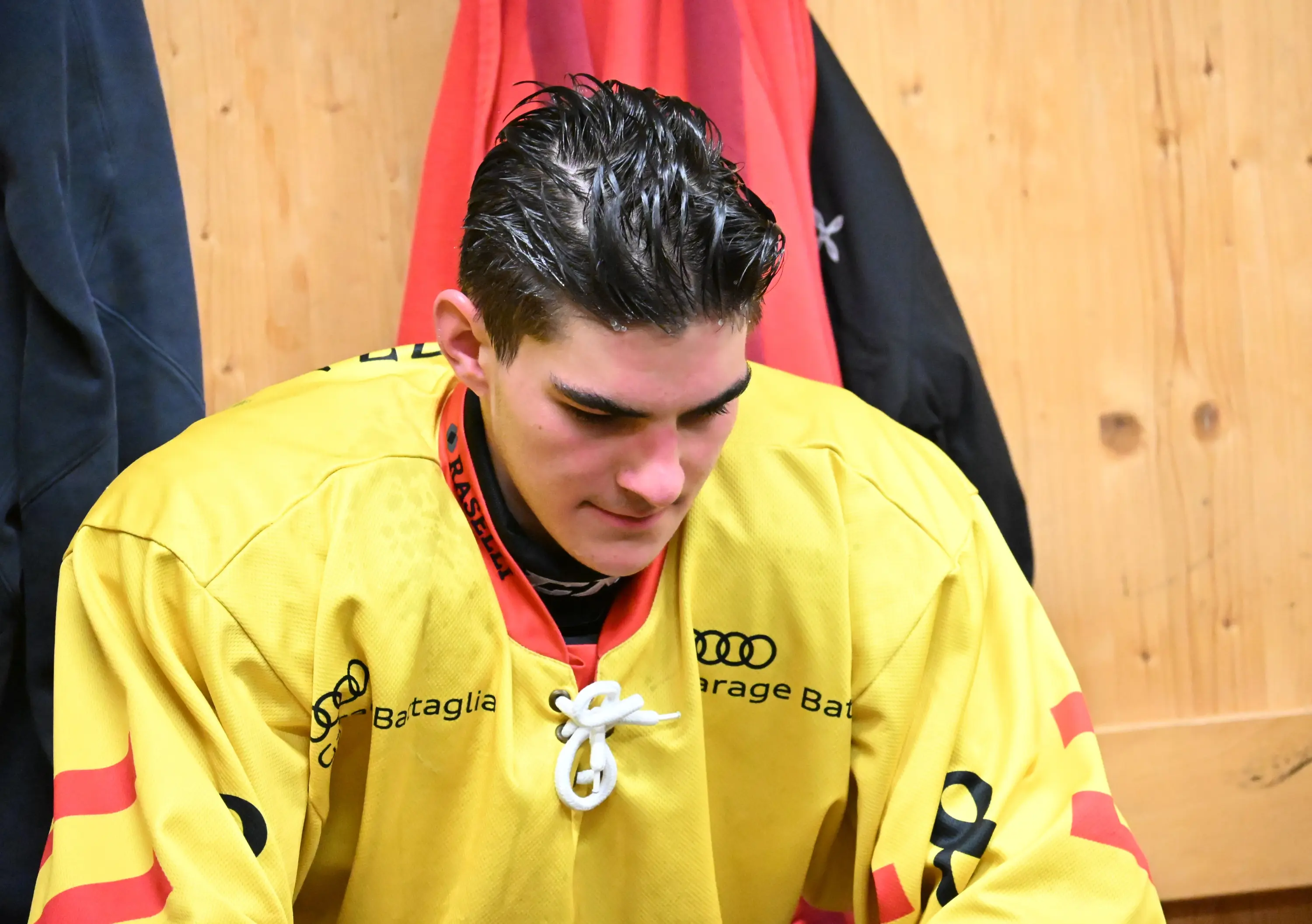 Giocatore di hockey su ghiaccio in divisa gialla, con capelli bagnati, si concentra all'interno di uno spogliatoio. Lo sfondo è di legno chi