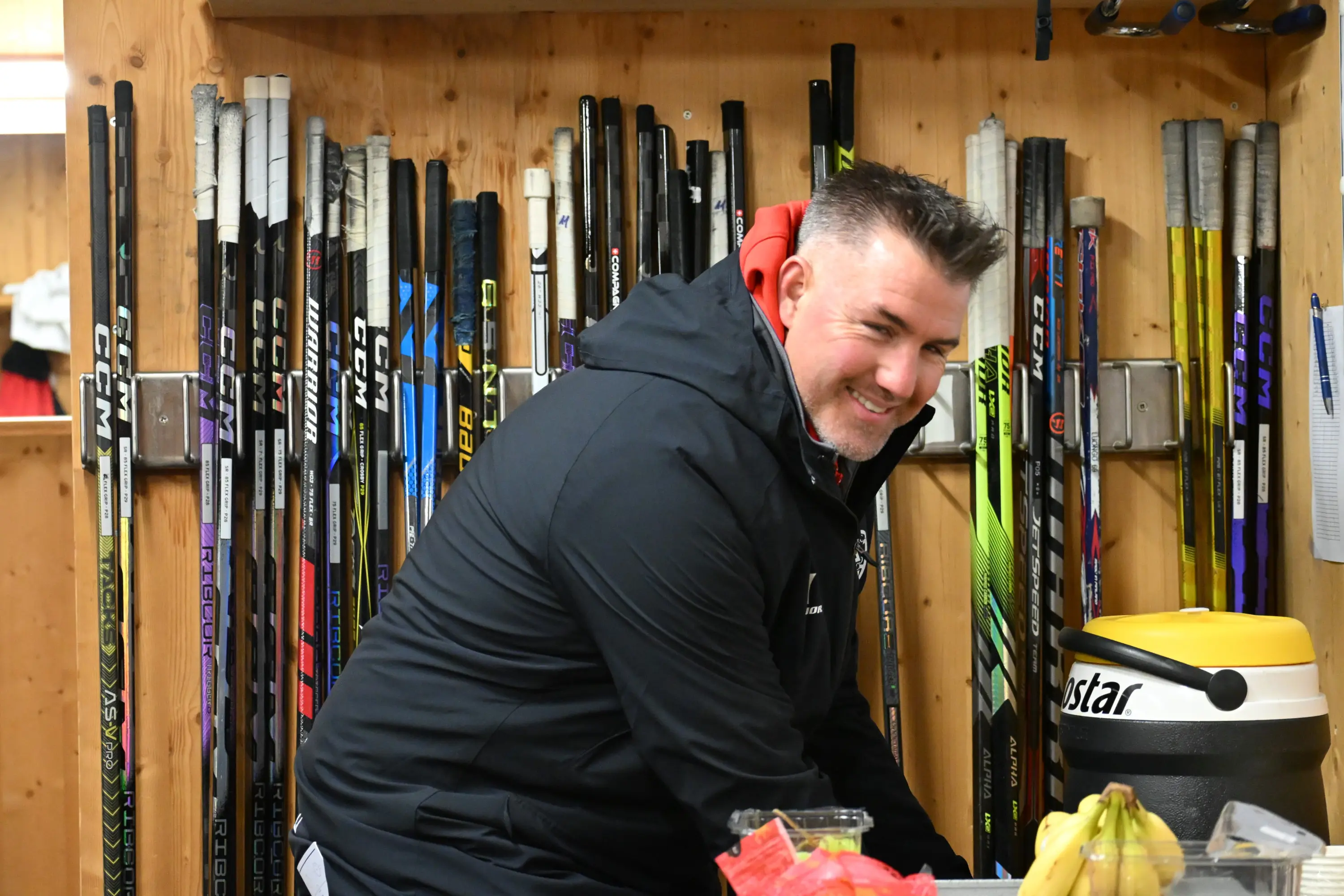 Un uomo sorridente indossa una giacca nera mentre si trova in un ambiente di spogliatoio, circondato da bastoni da hockey appoggiati su una parete di leg