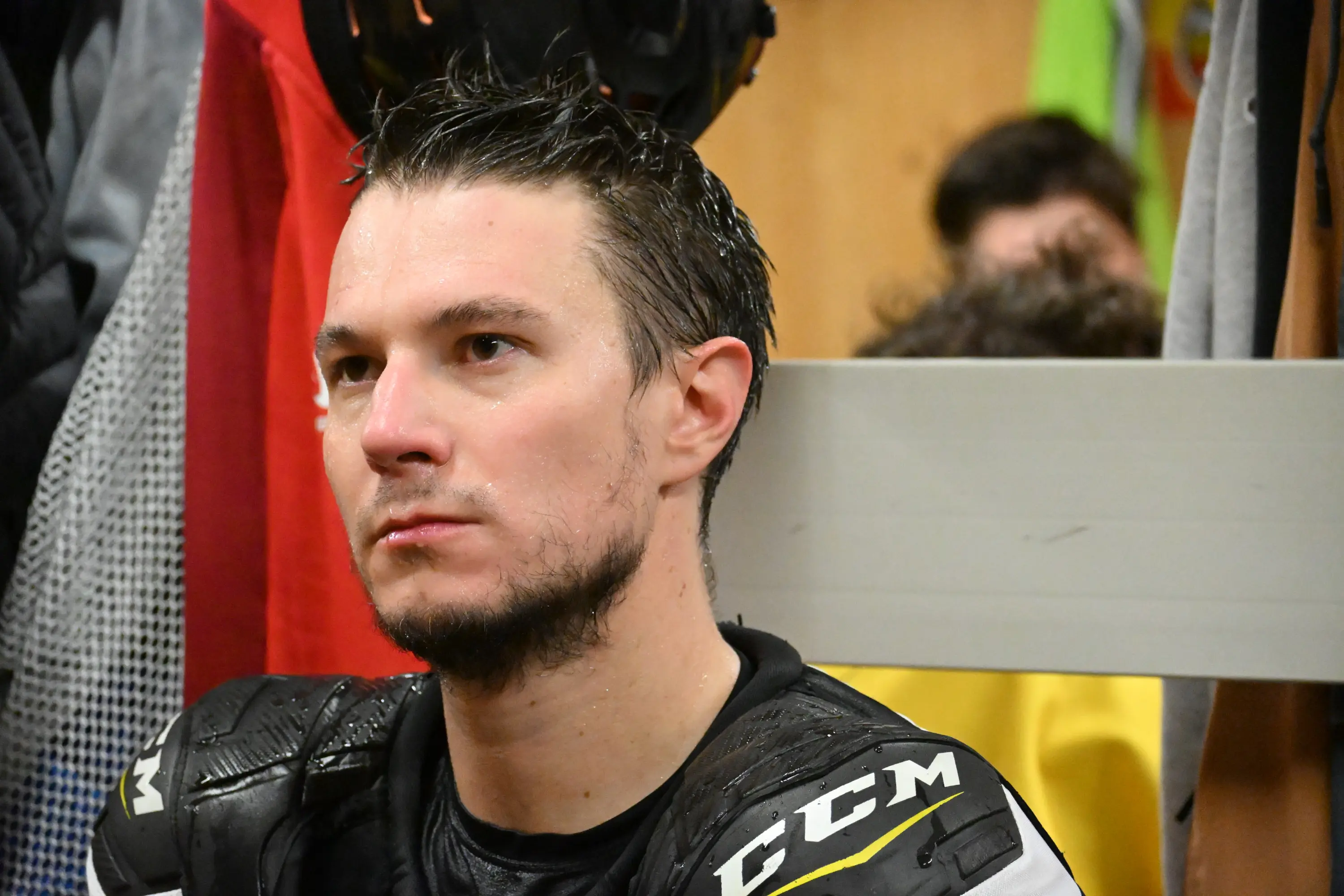 Giocatore di hockey con capelli bagnati e sguardo serio, seduto in uno spogliatoio. Indossa un'armatura da hockey e si intravedono giac