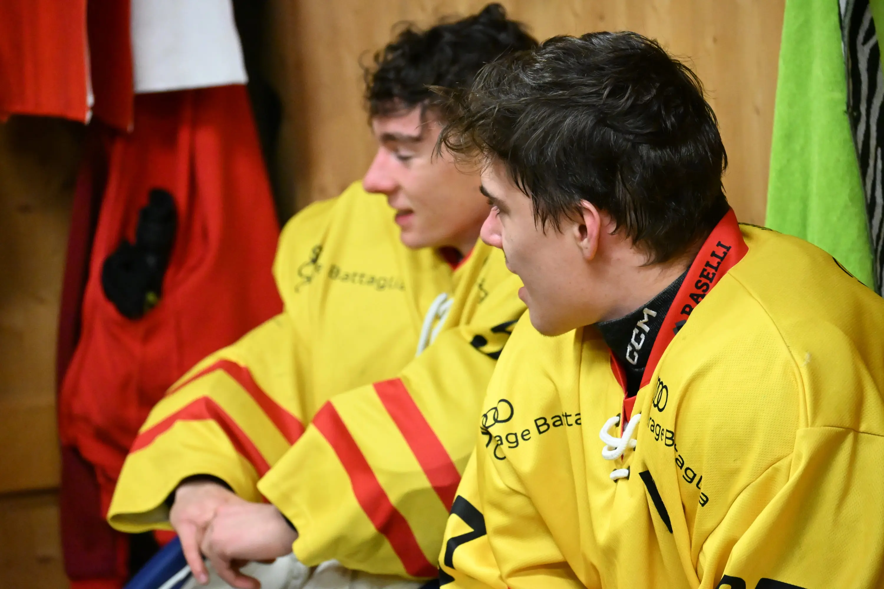 Due giovani atleti in divisa gialla, seduti in uno spogliatoio, sembrano conversare. Sullo sfondo si vedono alcune attrezzature sportive.