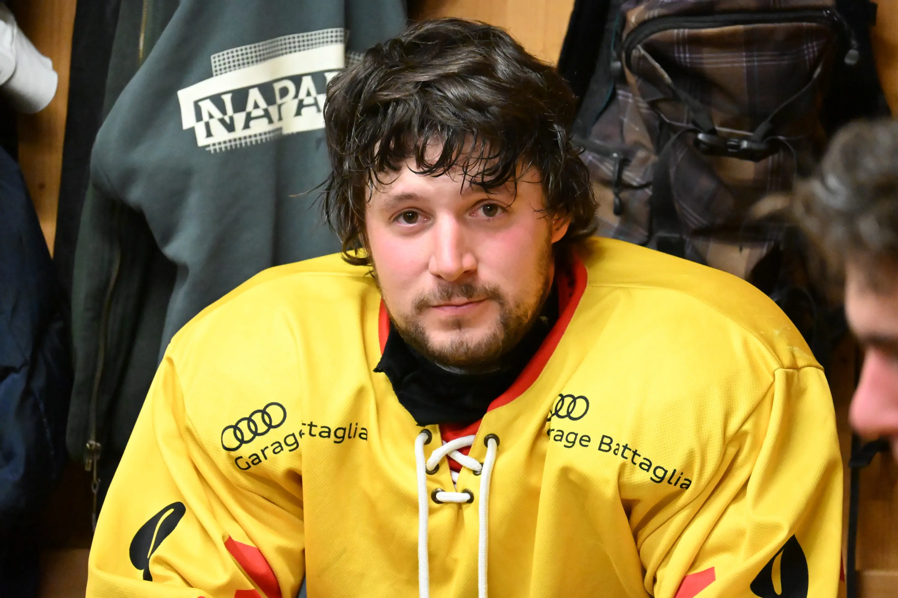 Giocatore di hockey su ghiaccio con capelli mossi, indossa una divisa gialla. Si trova in uno spogliatoio, in primo piano, con un'espressione seria