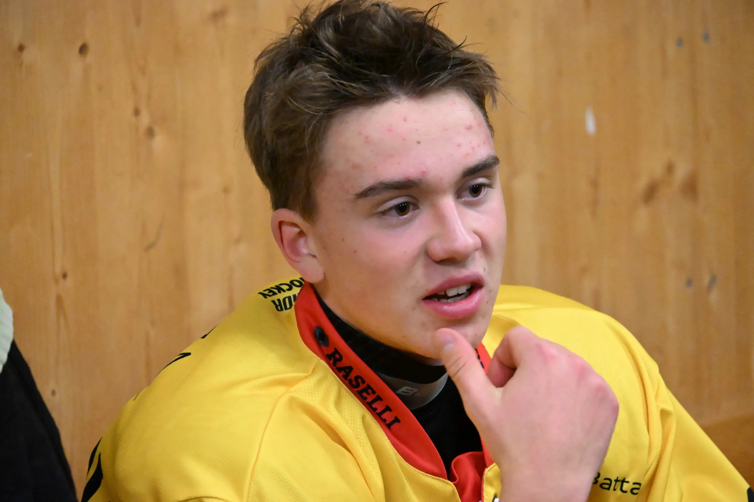 Giovane atleta seduto, indossa un giacca gialla e sembra riflettere. Sullo sfondo, una parete di legno. Mostra espressione concentrata.