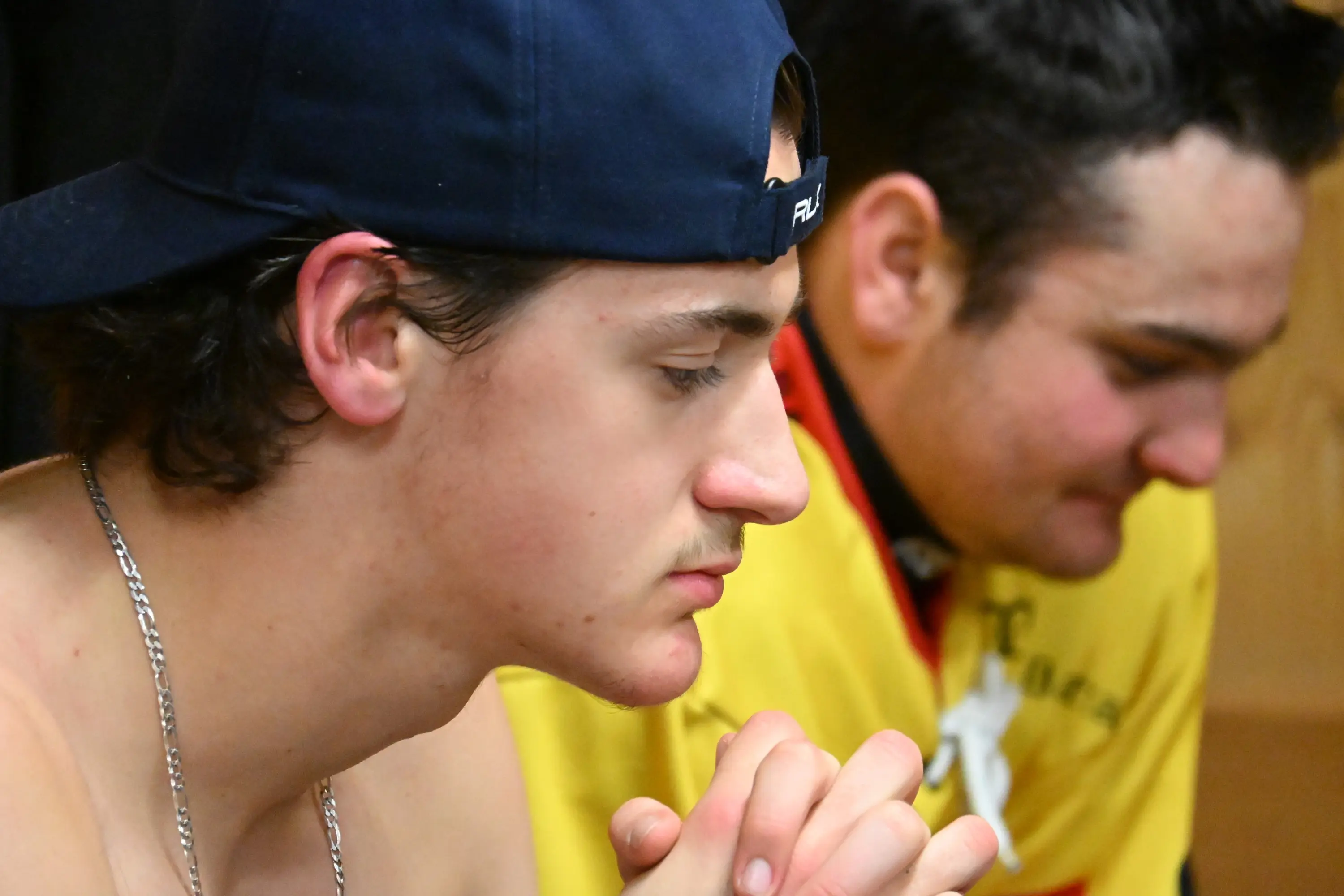 Due giovani uomini seduti, uno con un cappellino e catena al collo, l'altro in maglietta gialla, entrambi sembrano riflettere profondamente.