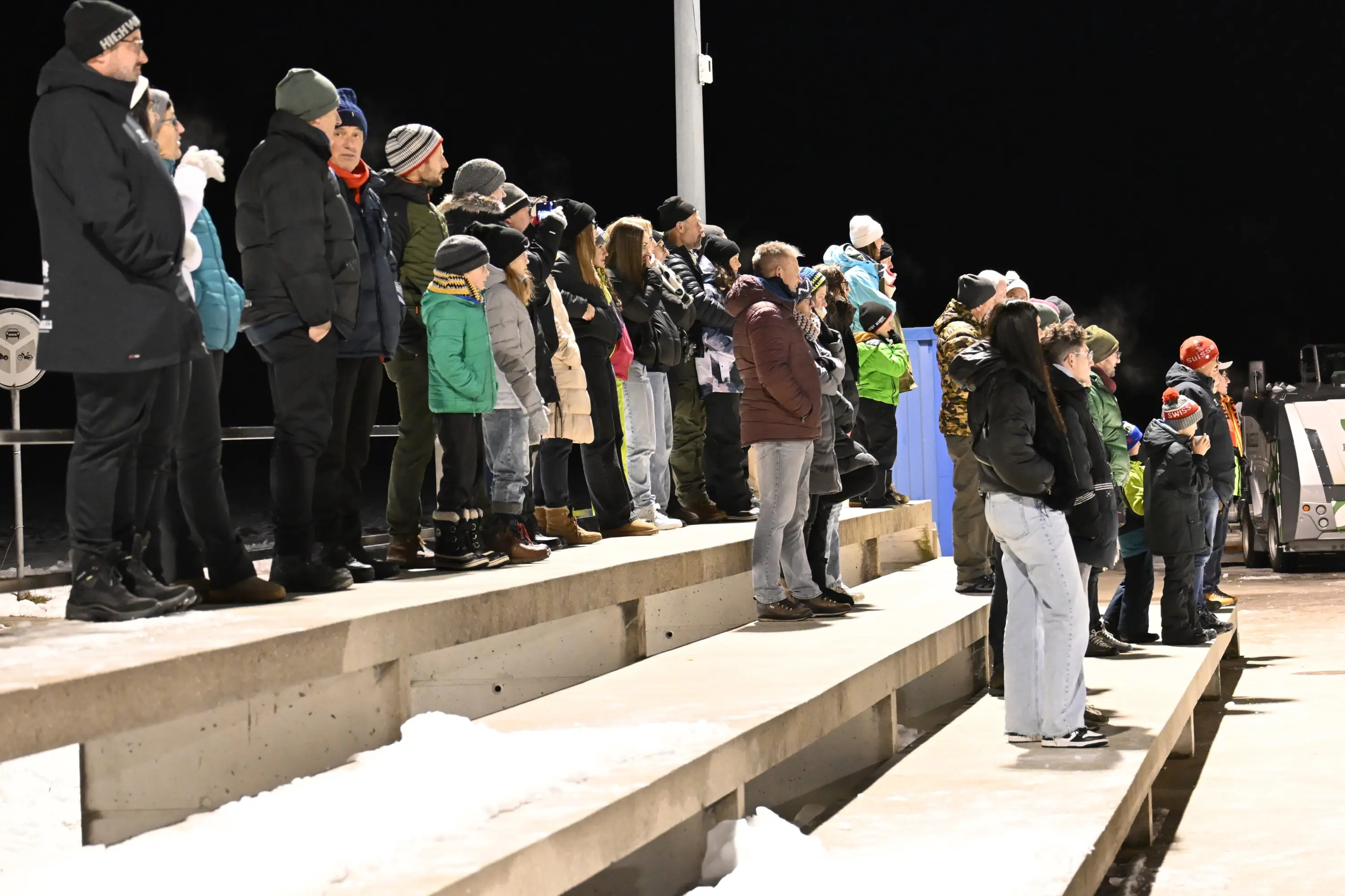 Un gruppo di persone in piedi su gradinate di cemento, vestite per il freddo, osservano un evento all'aperto di notte. Alcuni indossano cappelli e giac