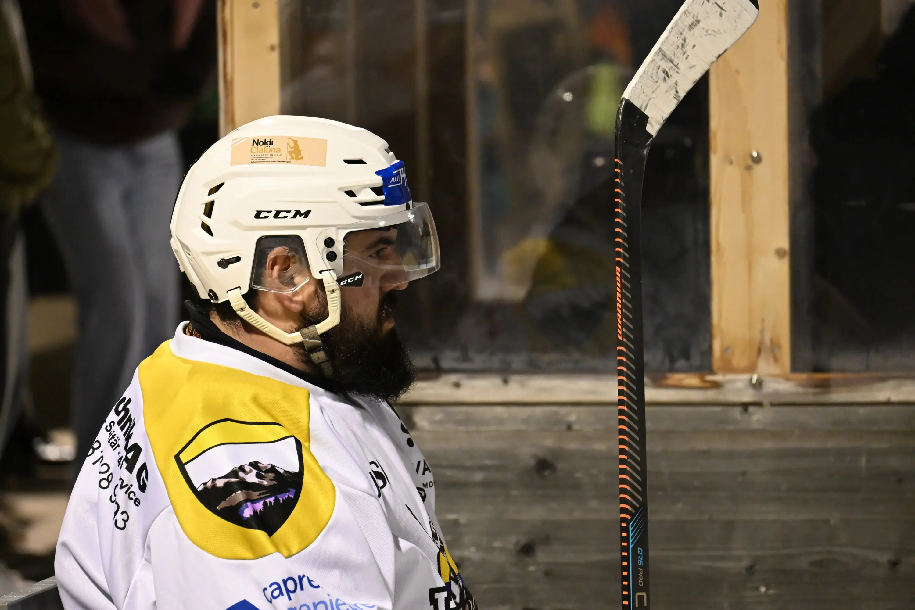 Giocatore di hockey con casco bianco, protezioni e bastone da hockey seduto in panchina, concentrato sul gioco.