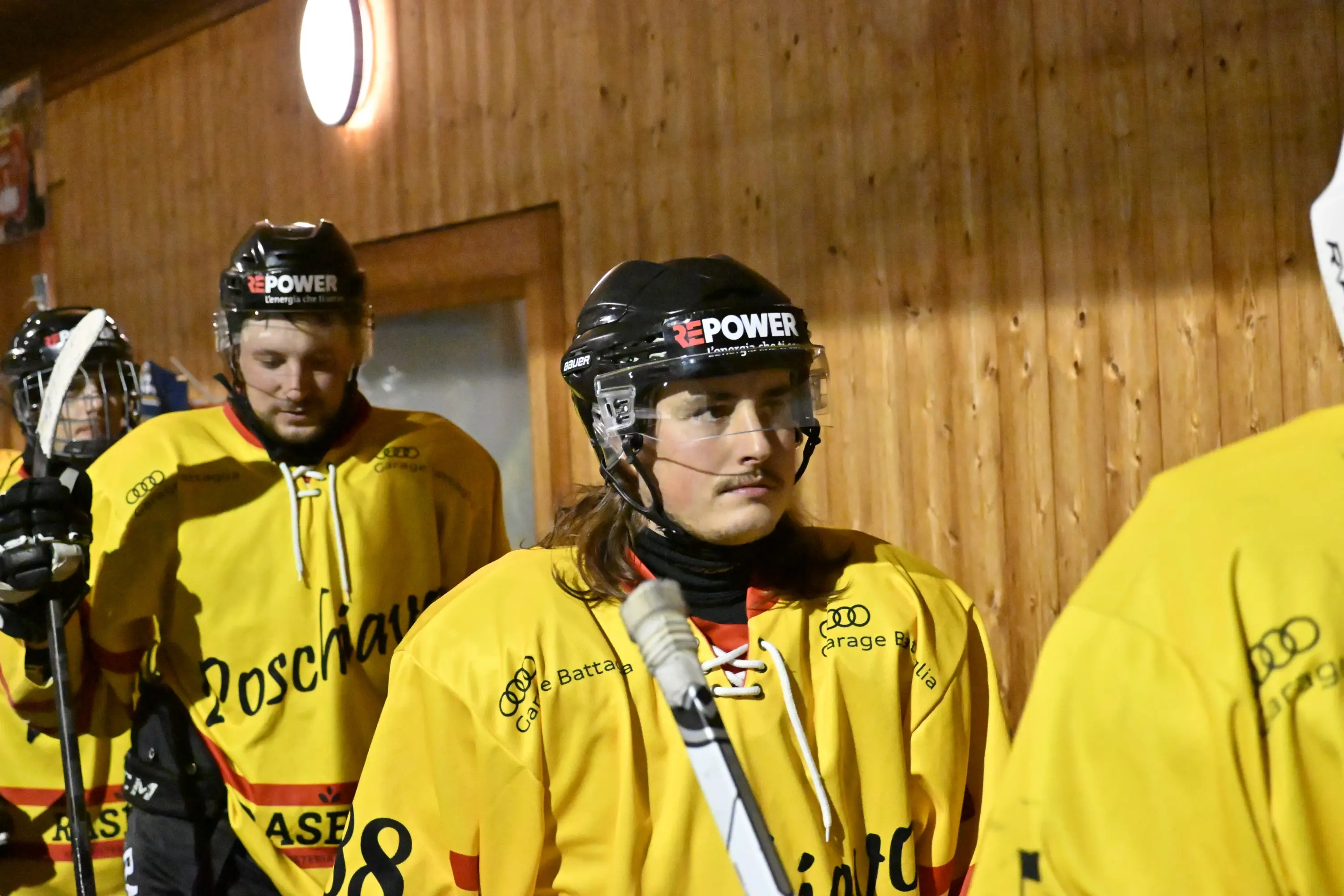 Giocatori di hockey su ghiaccio in caschi e maglie gialle si preparano per entrare in campo. Uno dei giocatori, con lunghi capelli, sembra concentrato.
