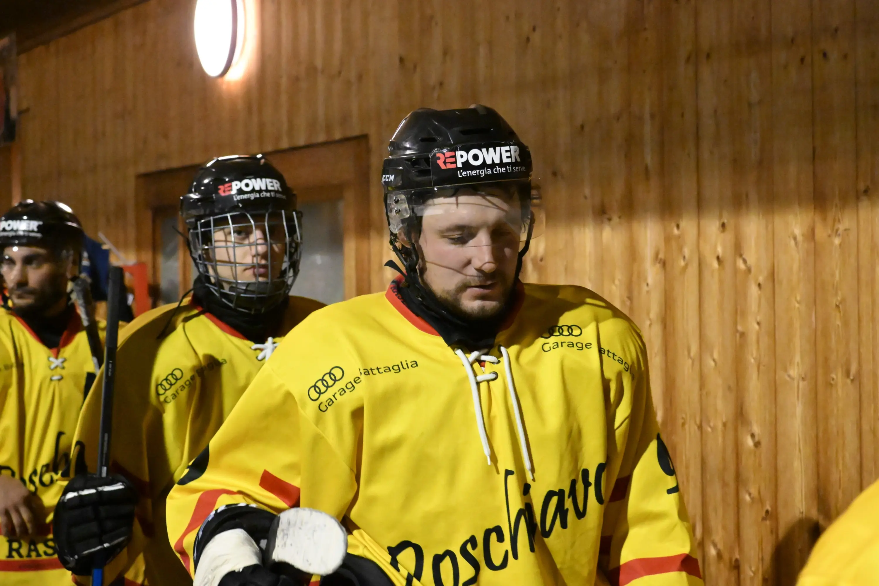 Giocatori di hockey su ghiaccio in divisa gialla, con elmetti, che si preparano a entrare in campo. Lo sfondo è in legno, creando un'at