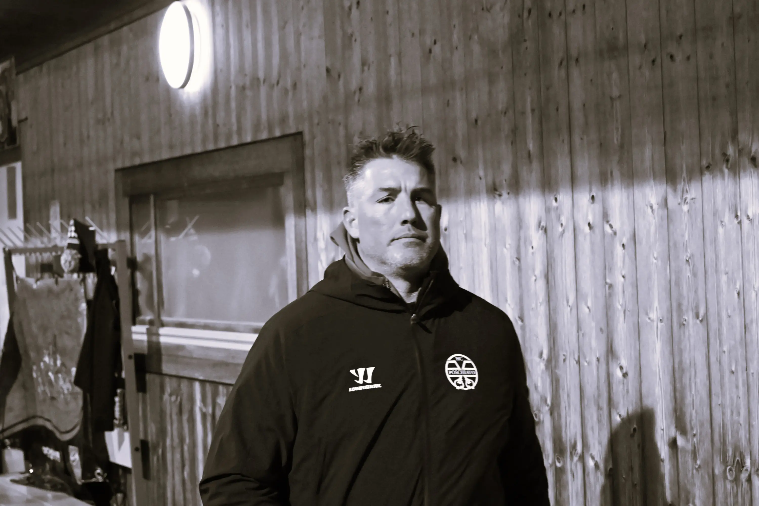 Uomo con giacca scura e capelli grigi, in piedi accanto a una parete di legno, con una luce circolare sopra di lui. Il suo sguardo è serio