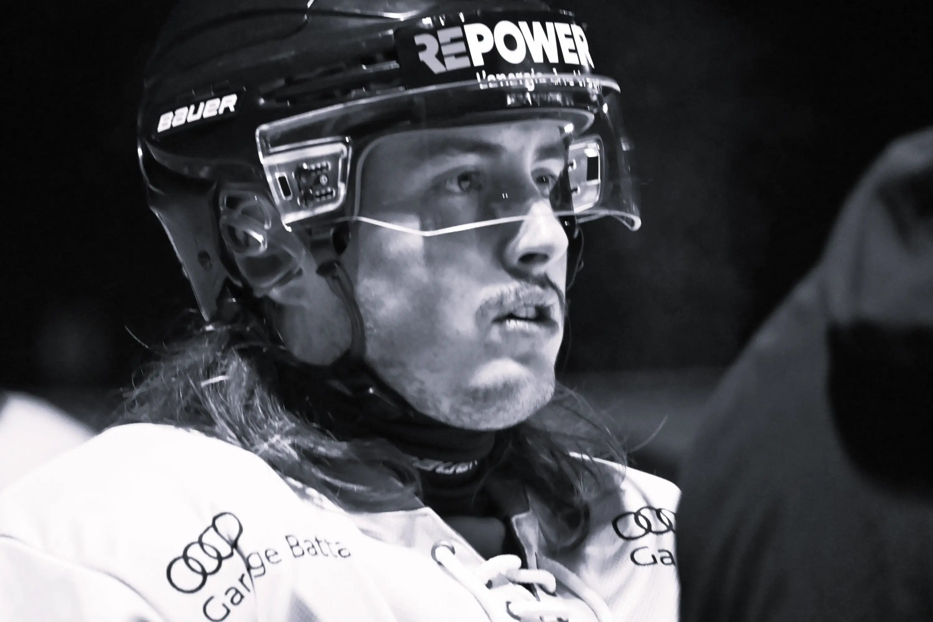 Giocatore di hockey su ghiaccio in uno momento di concentrazione, con capelli lunghi e un baffo, indossa un casco e una maglia da gioco.