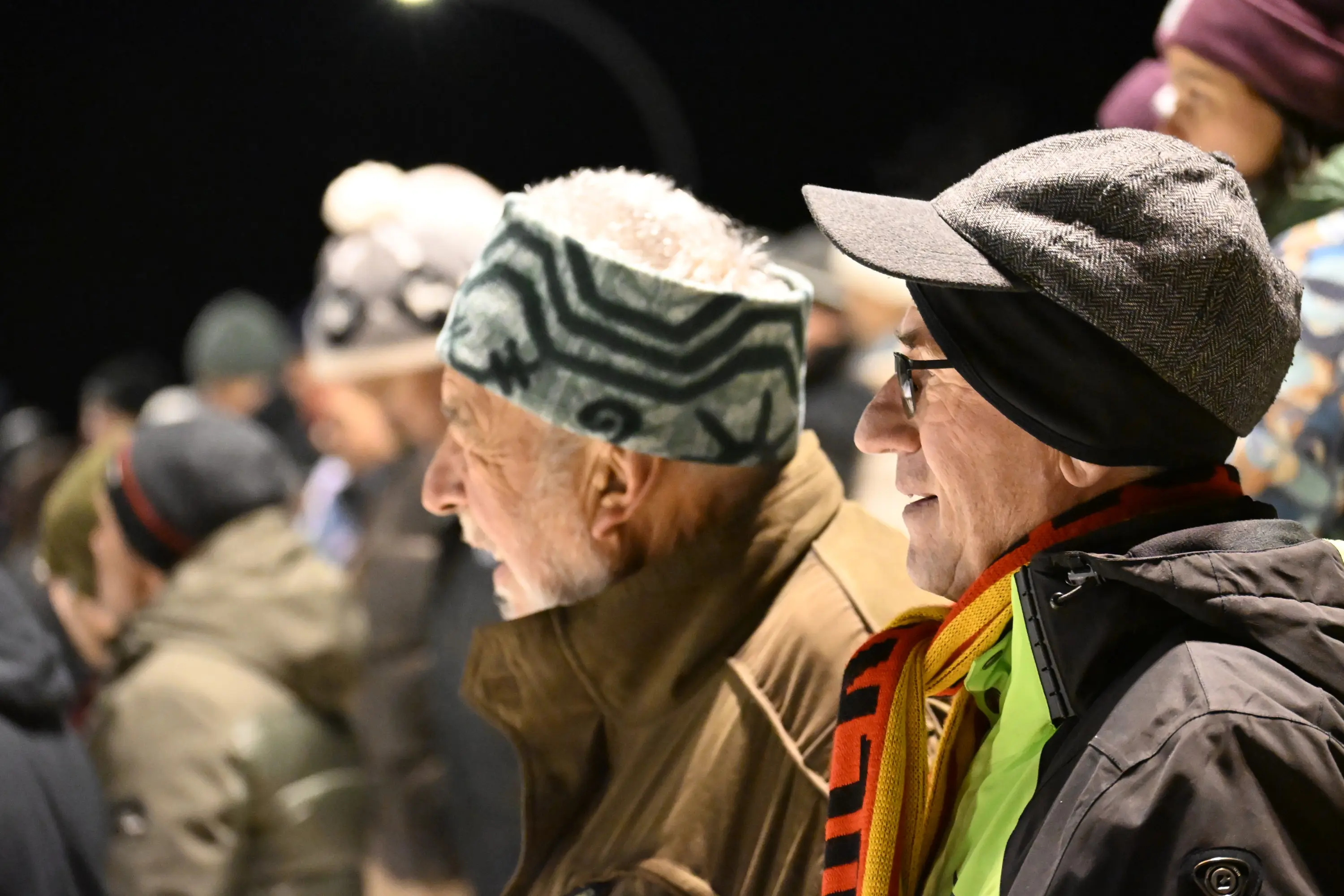 Due uomini anziani osservano attentamente, indossando berrettini e sciarpe colorate. Sullo sfondo si intravede un pubblico entusiasta. È notte e ci