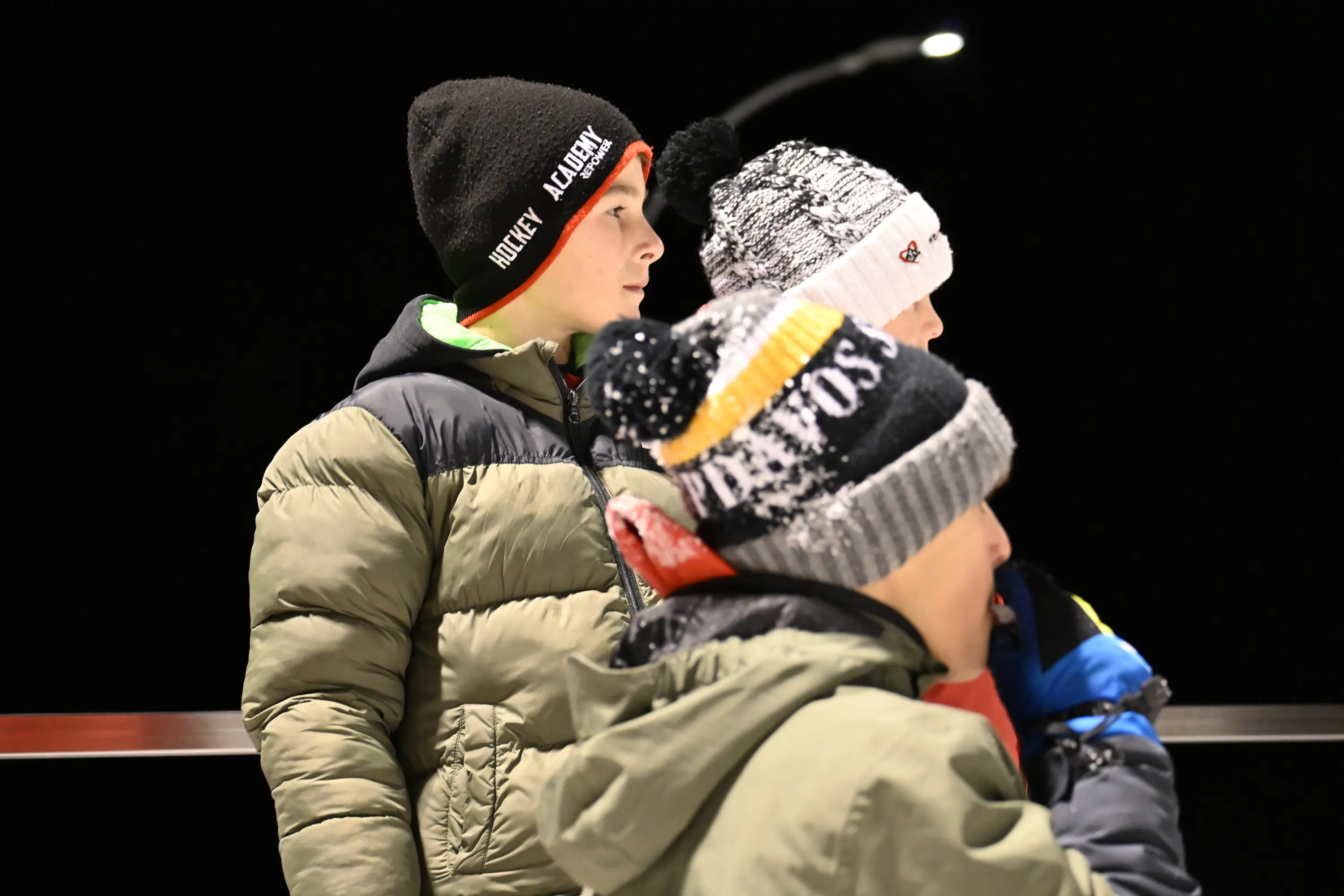 Tre bambini indossano berretti invernali e giacche, osservando qualcosa in lontananza. La scena è illuminata da una luce notturna.