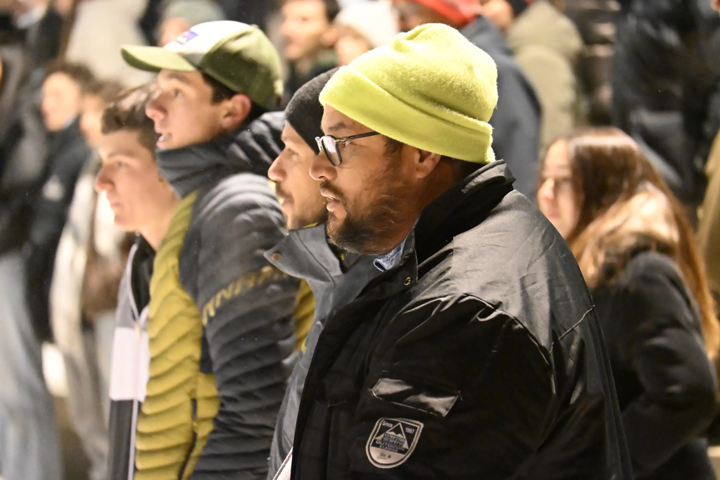 Gruppo di persone in un evento all'aperto, con un uomo in primo piano che indossa un cappello giallo e una giacca nera. Sullo sfondo si intraved