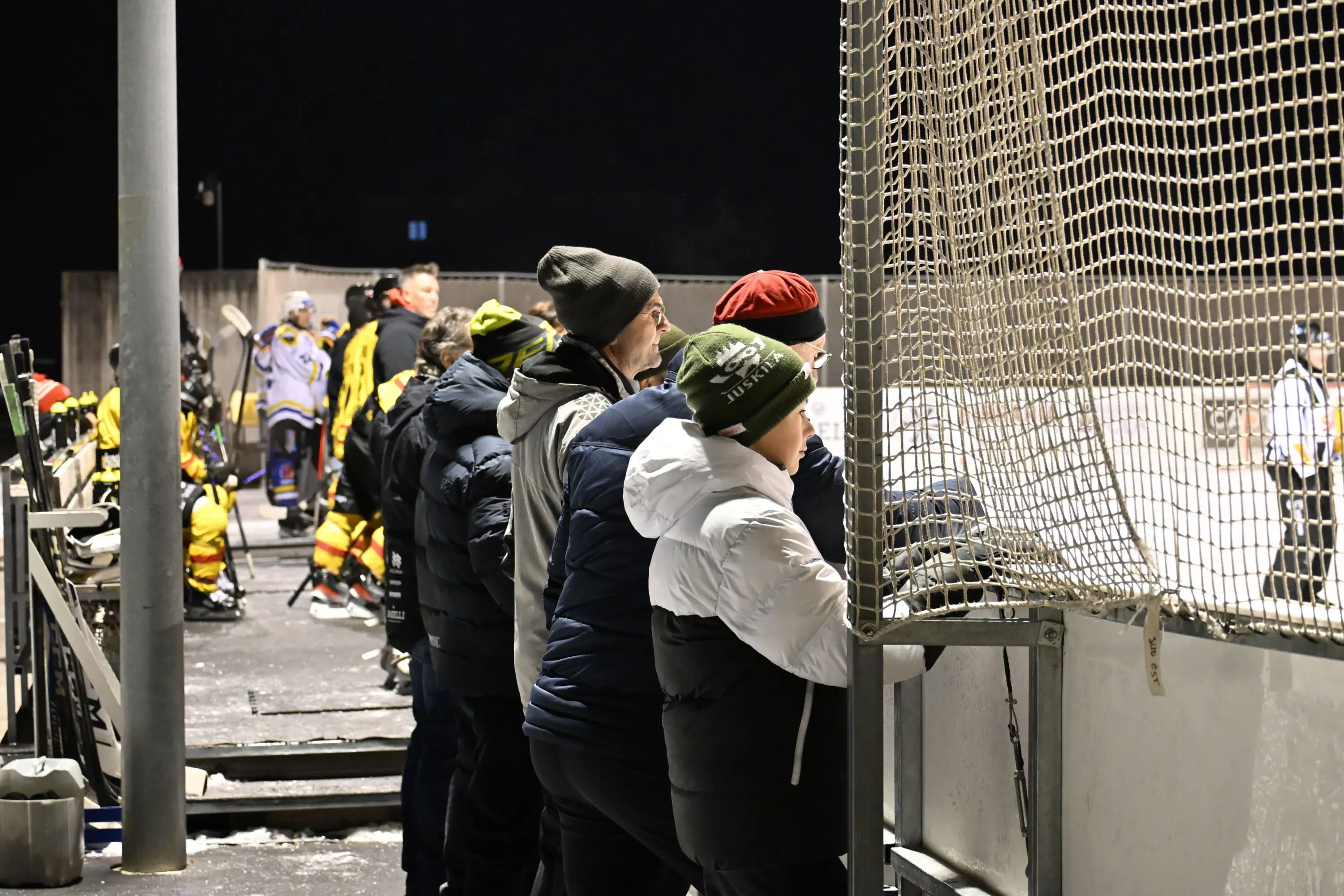 Un gruppo di persone osserva una partita di hockey su ghiaccio da dietro una rete. Alcuni indossano giacche calde e berretti, mentre sullo sfondo ci sono