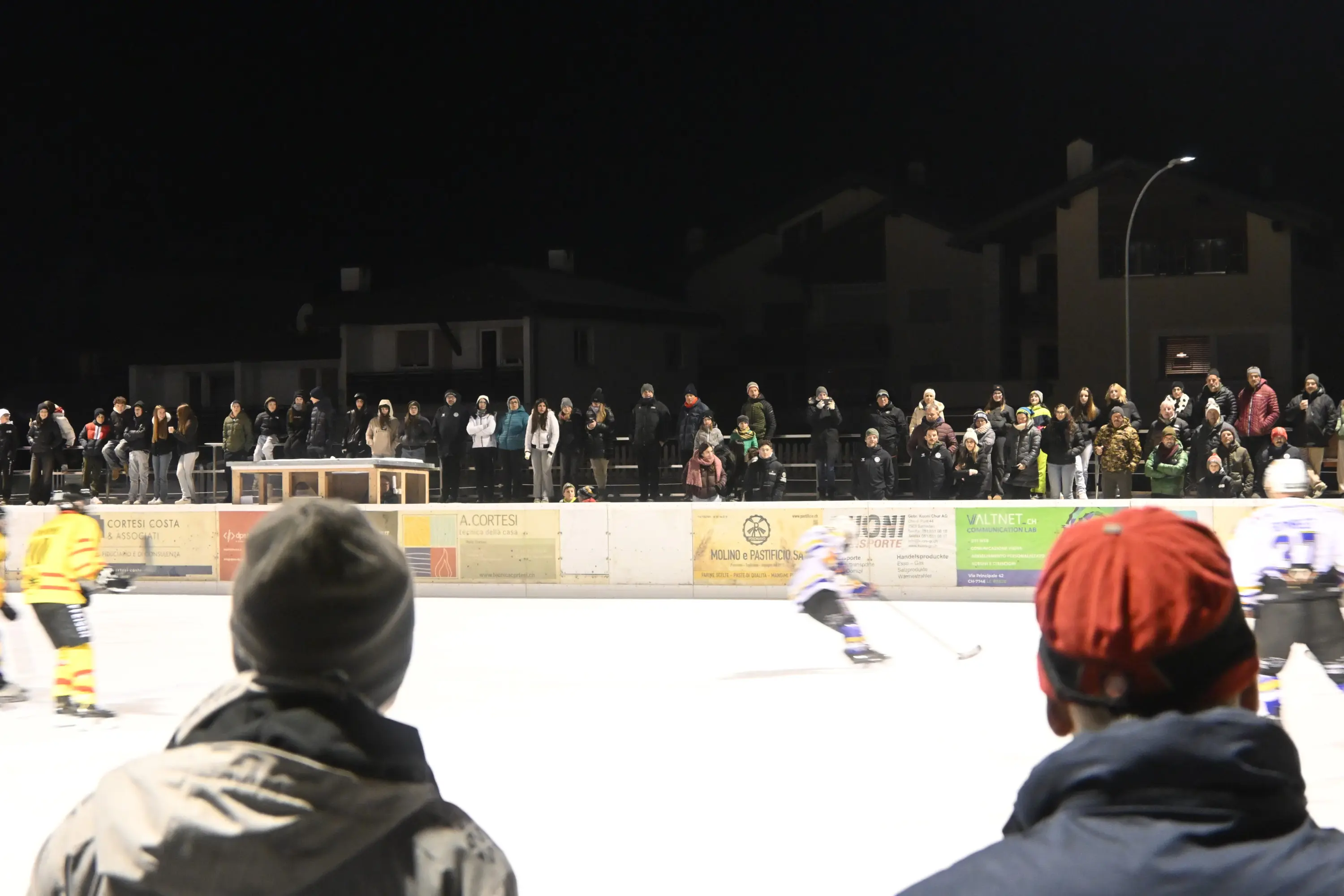 Un gruppo di persone assiste a una partita di hockey su ghiaccio all'aperto di notte. La scena è affollata, con spettatori che si sono radunati lungo le balaustre