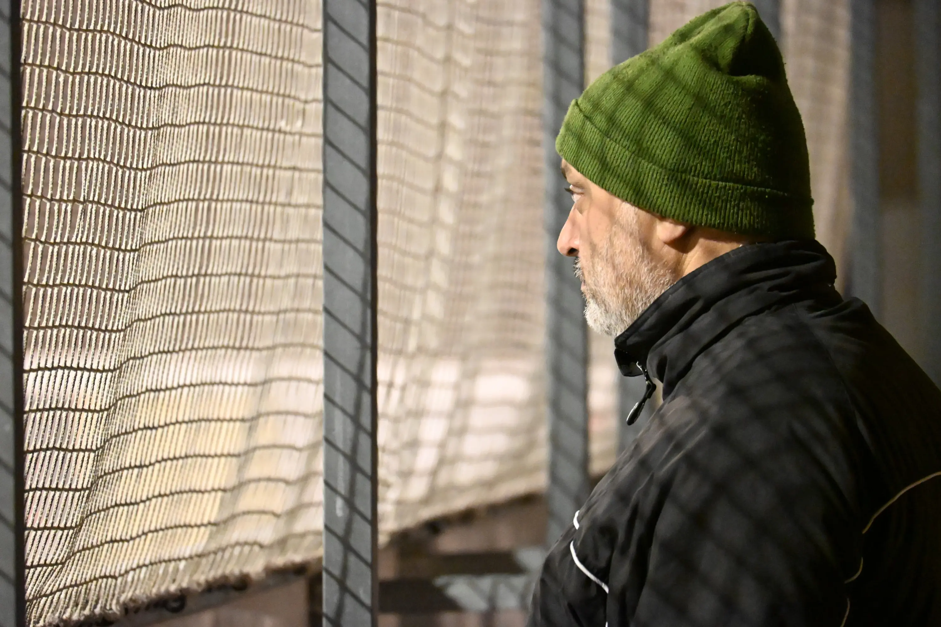 Un uomo con una berretto verde osserva attraverso una rete metallica. Indossa un giubbotto nero e ha i capelli brizzolati.