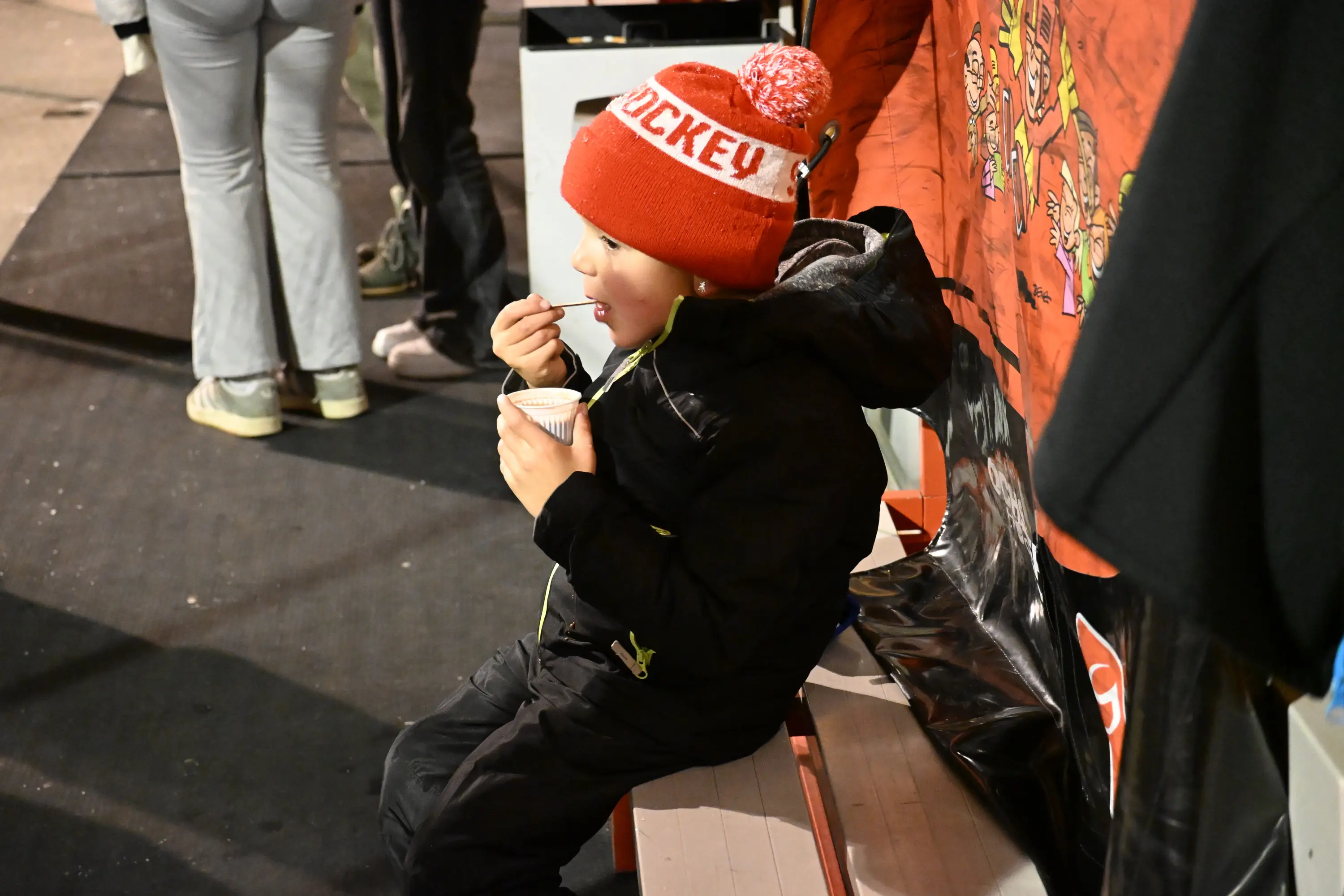 Un bambino seduto su una panchina indossa un berretto rosso con la scritta "Hockey". Sta mangiando un gelato e guarda la scena attorno a lui.