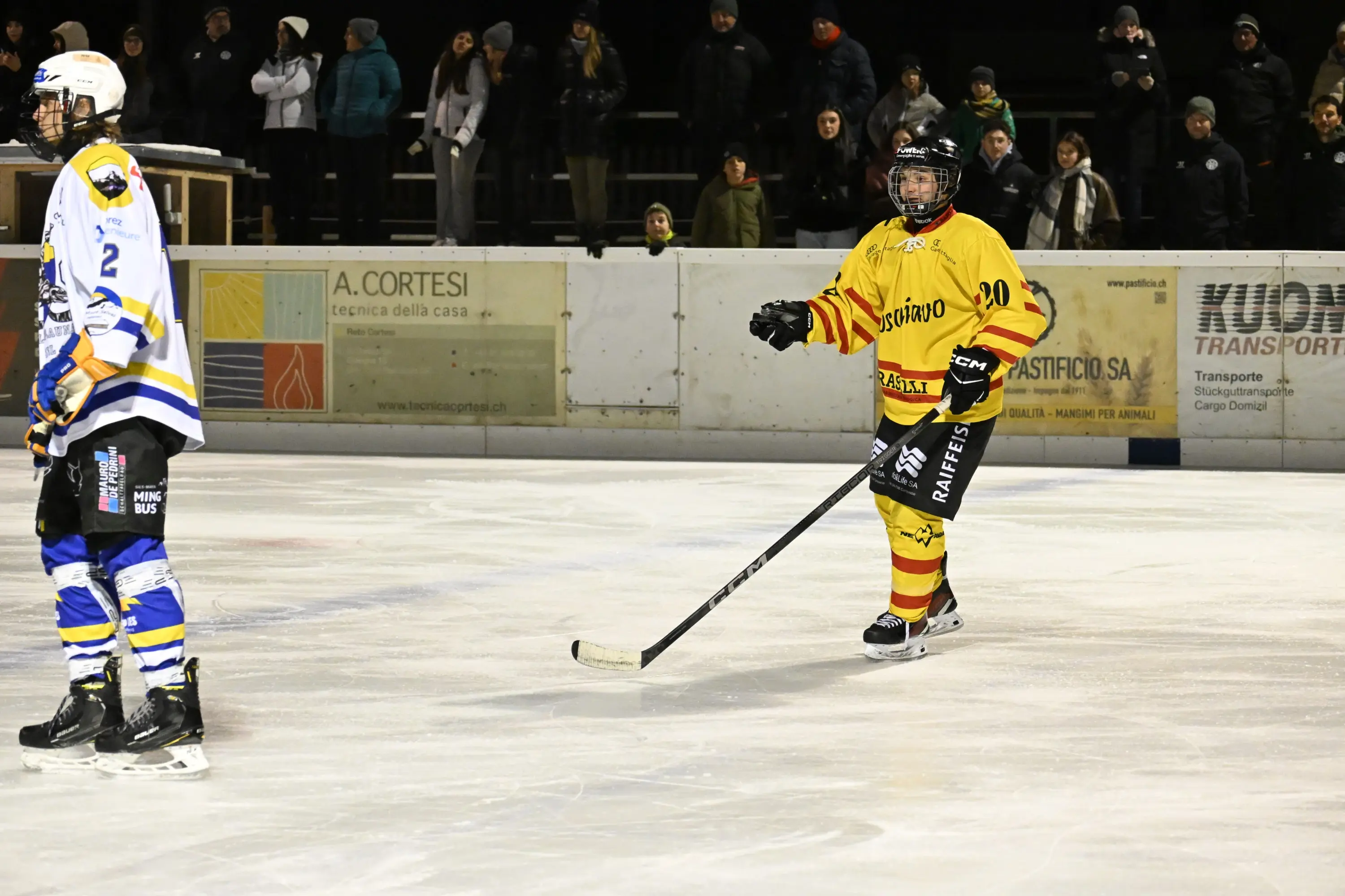 Giocatori di hockey su ghiaccio in un campo, uno indossa una maglia gialla con strisce rosse, l'altro una maglia blu e bianca. Sullo sf