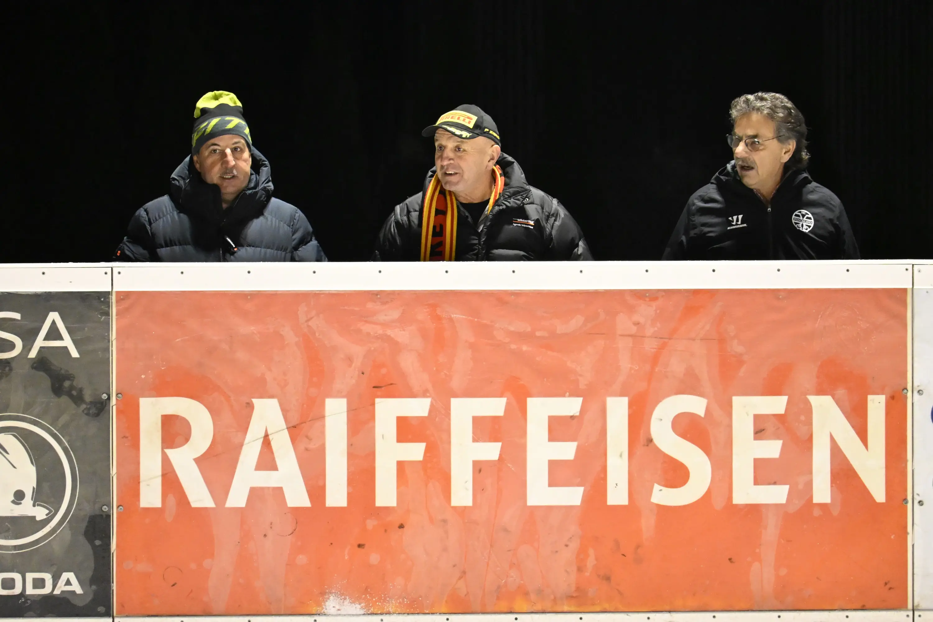 Tre uomini sorridenti stanno seduti dietro un pannello pubblicitario rosso con la scritta "RAIFFEISEN". Indossano abbigliamento invernale e si trov