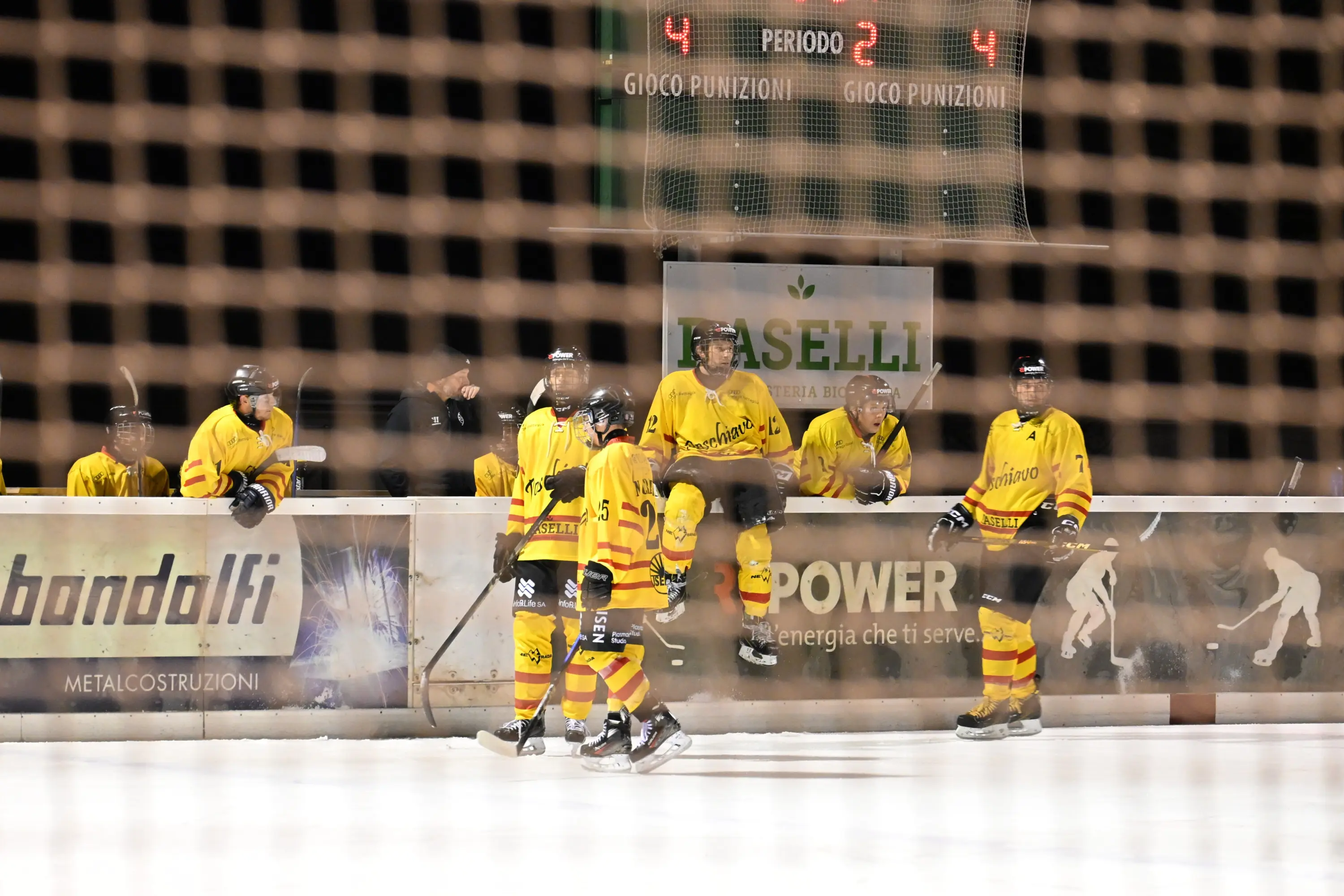 Giocatori di hockey su ghiaccio in divisa gialla siedono e si preparano sulla panchina durante una partita. Il punteggio è 4-2.