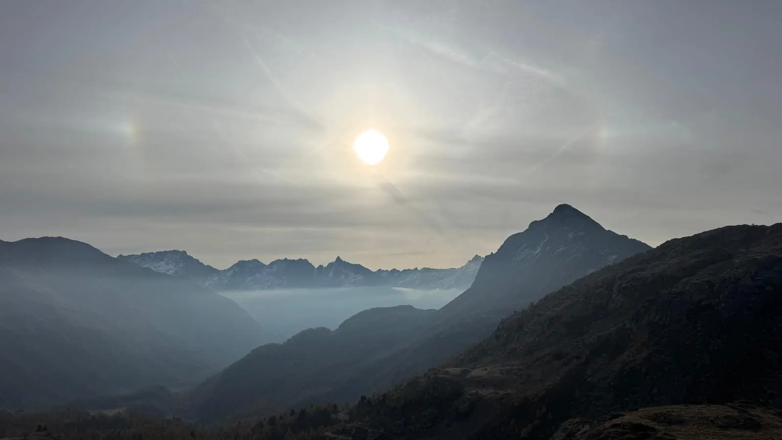 Paesaggio montuoso al tramonto, con un sole luminoso e aloni solari visibili. Le montagne si stagliano all'orizzonte, avvolte da una leggera foschia.