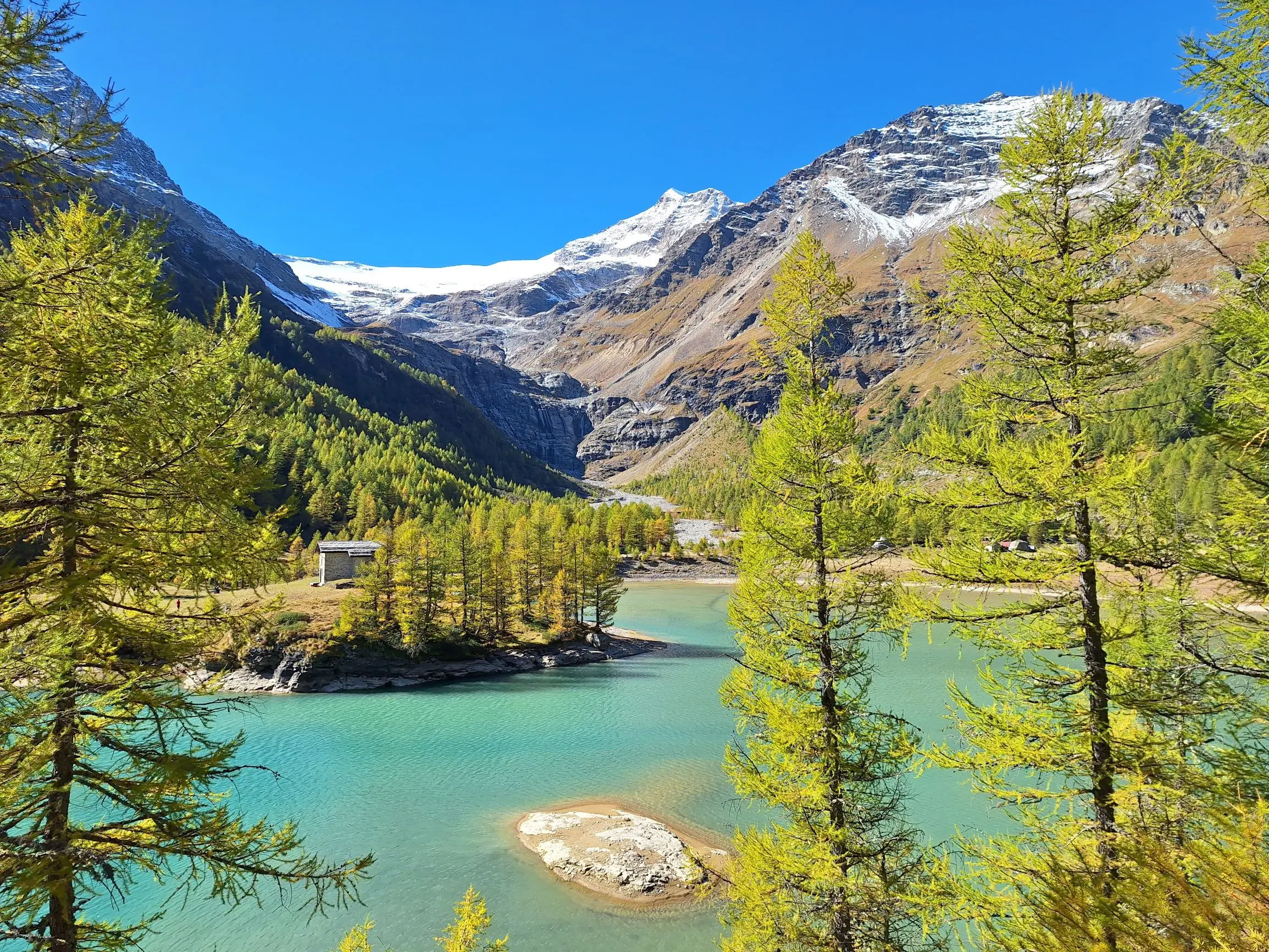 Una vista panoramica di un lago turchese circondato da alberi verdi. Sullo sfondo, imponenti montagne con cime innevate e un cielo blu. Un'isoletta sabbiosa si trova al centro del lago.