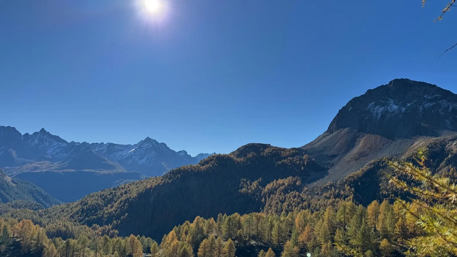Un ampio panorama montano con cime innevate e colline ricoperte di alberi autunnali. Il sole splende nel cielo blu, creando un'atmosfera serena.