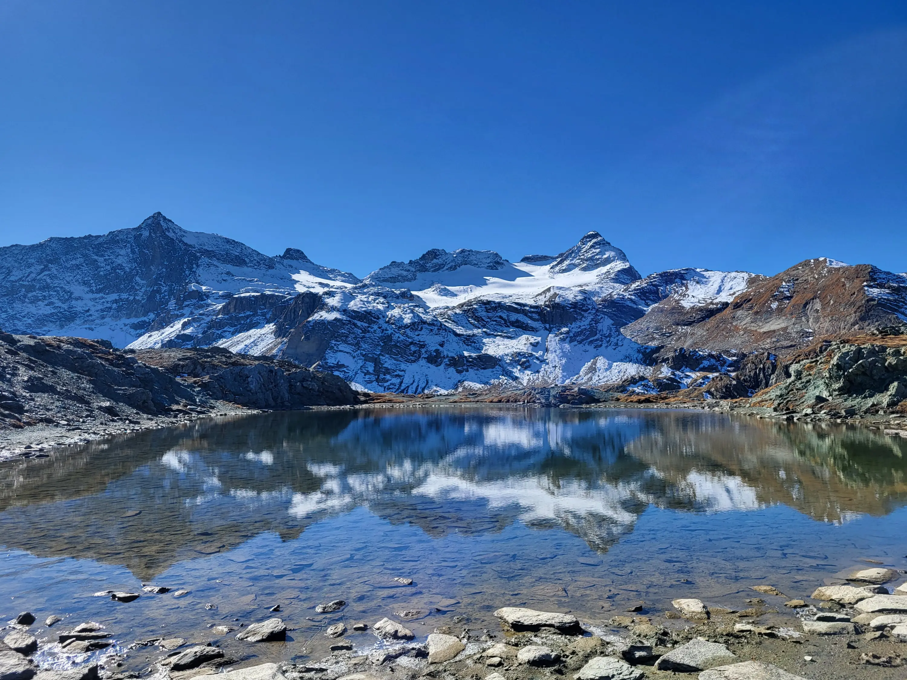 Un lago alpino cristallino riflette imponenti montagne coperte di neve e un cielo blu limpido. Rocce e ghiaia bordano il lago.