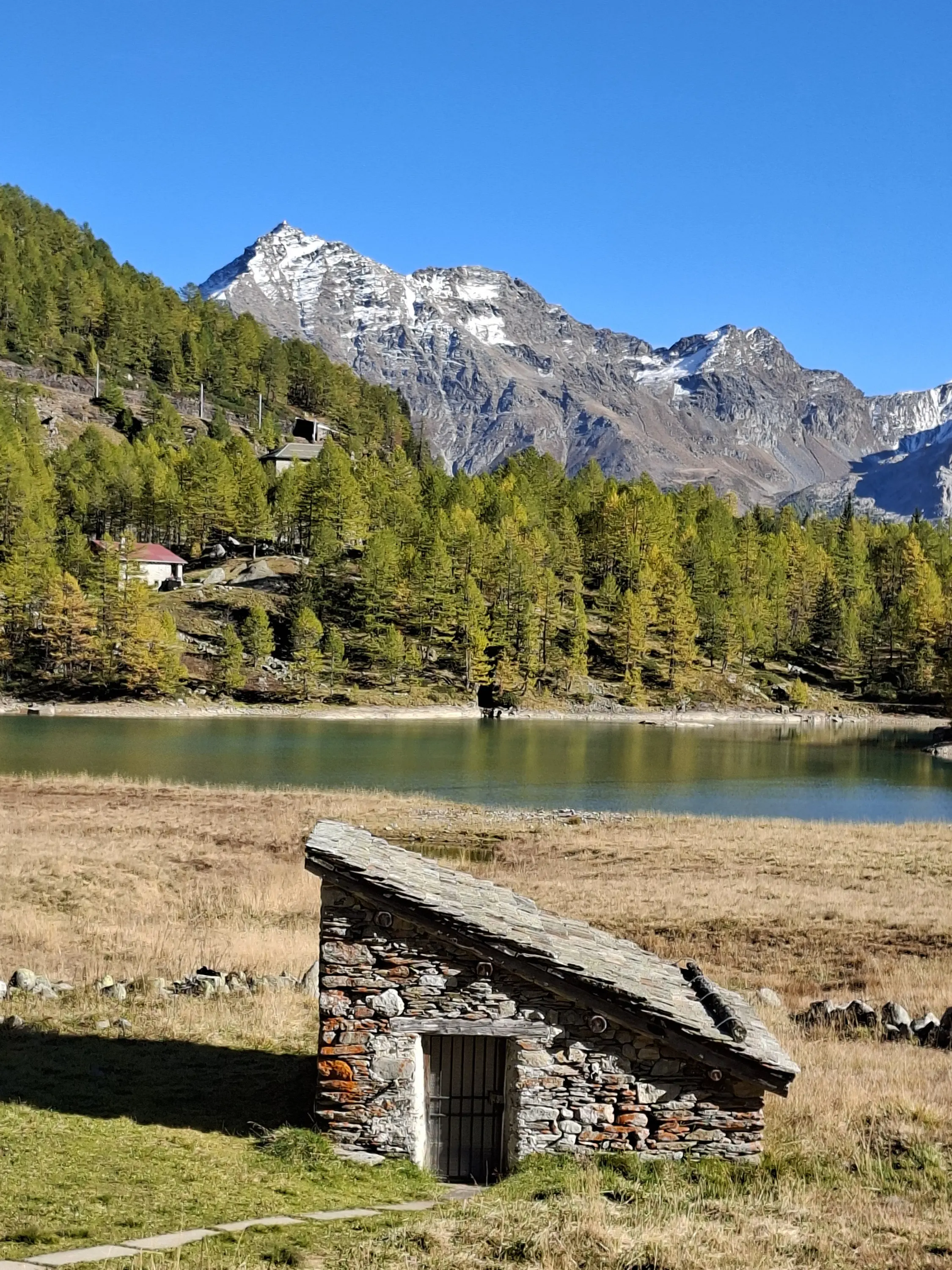 Un piccolo edificio in pietra con il tetto inclinato si trova in un prato vicino a un lago, circondato da alberi verdi e montagne innevate sullo sfondo, in una giornata di sole.