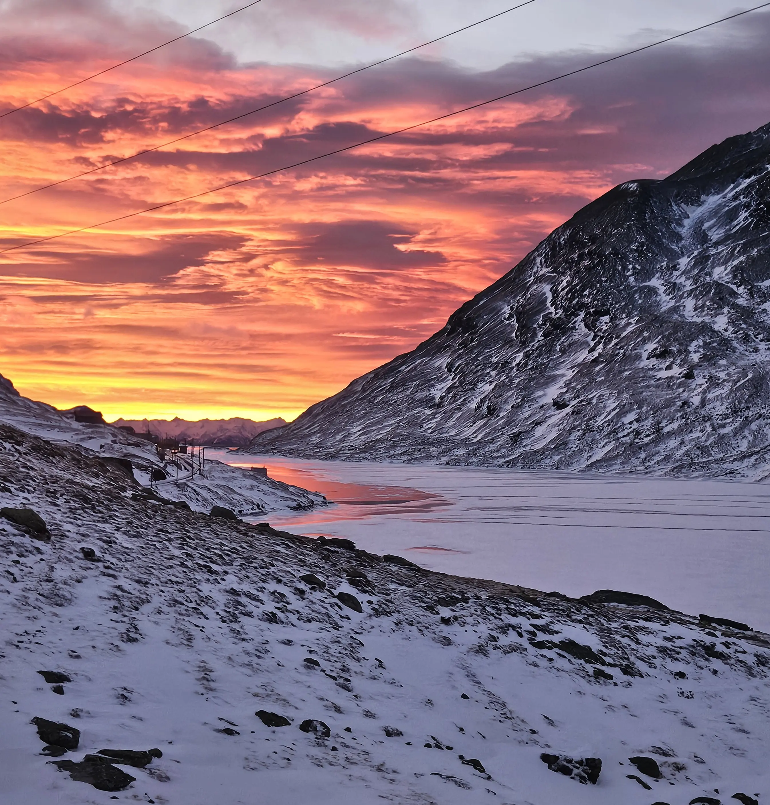 Un paesaggio montano al tramonto, con un cielo che sfuma rosso e arancione. La luce si riflette su un fiume ghiacciato, mentre le montagne circostanti sono coperte di neve.