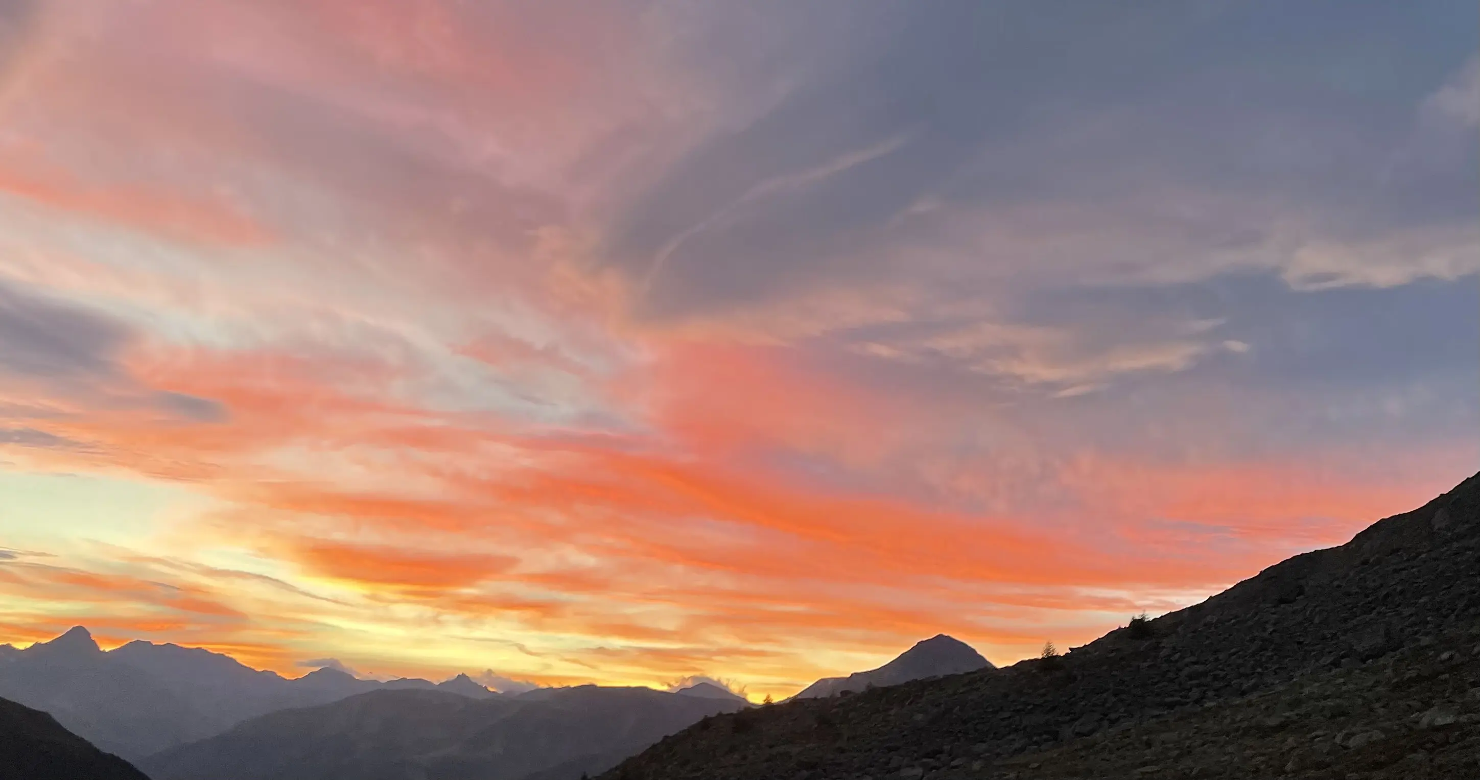 Un tramonto spettacolare dipinge il cielo con tonalità di arancione, rosa e blu, mentre silhouette montuose si stagliano in primo piano.