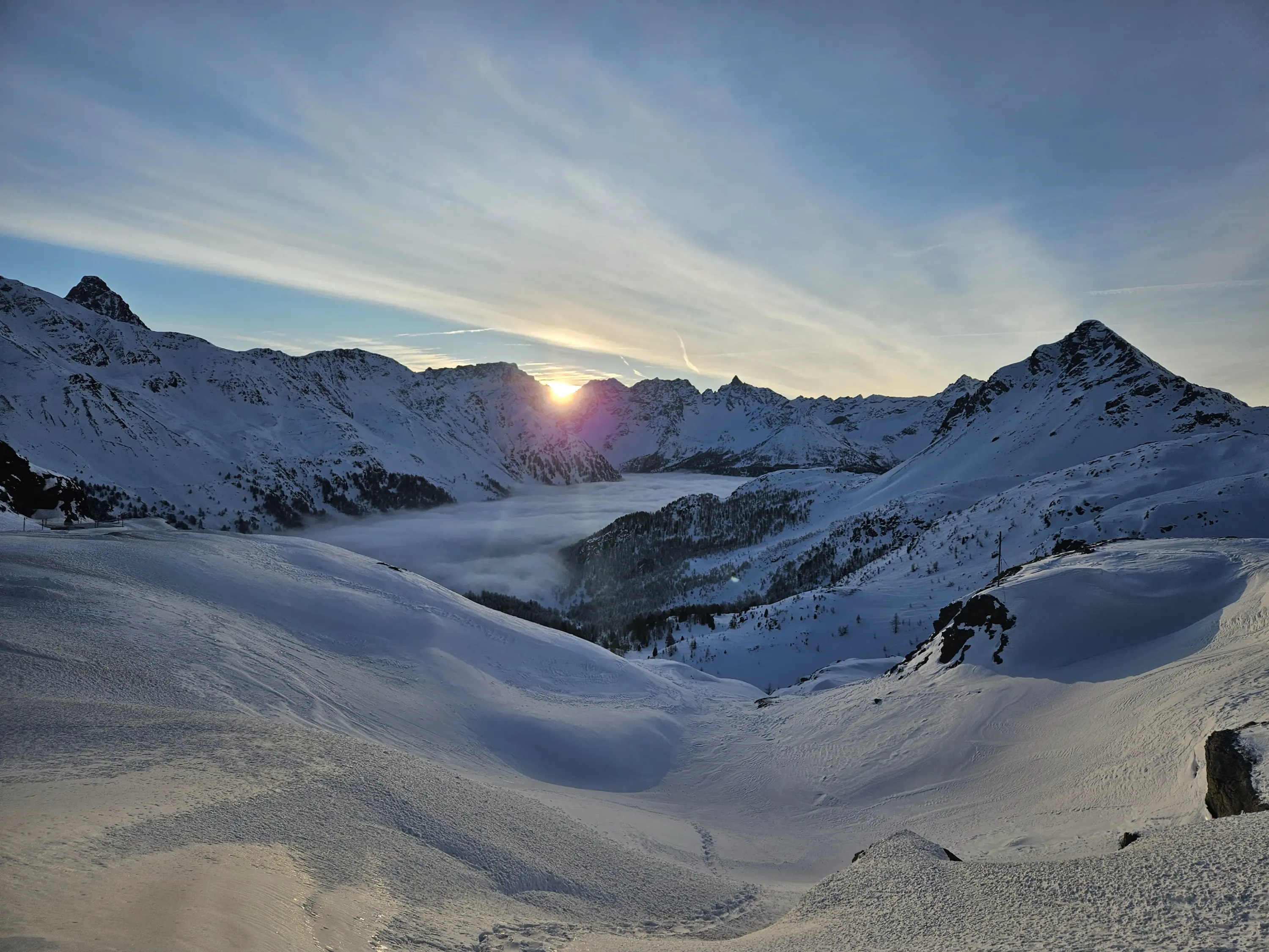 Un paesaggio montano innevato al tramonto, con vette illuminate dalla luce dorata e una valle avvolta nelle nuvole.