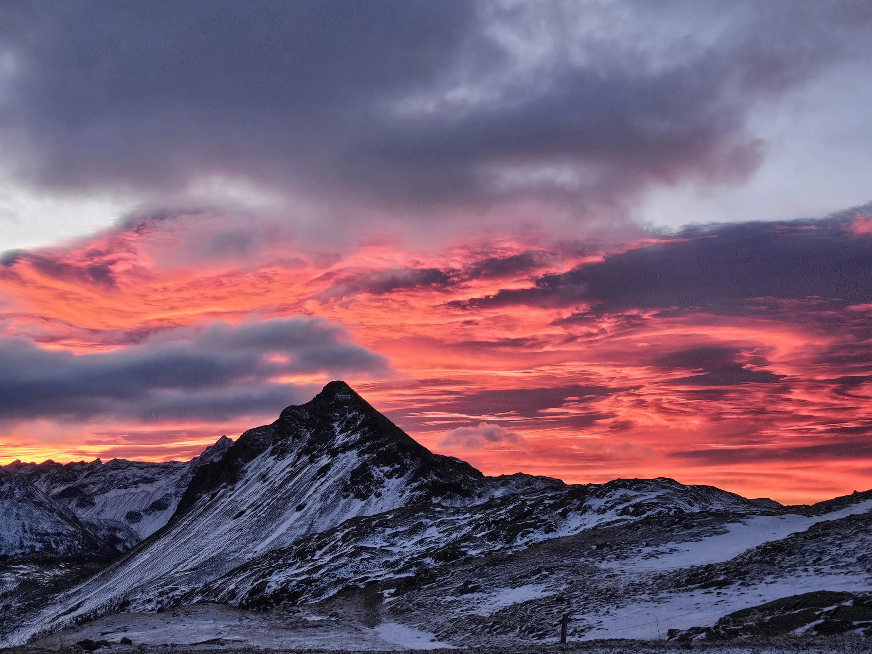 Un paesaggio montano al tramonto, con nuvole arancioni e rosa che si stagliano contro un cielo grigio. Un picco montano coperto di neve è visibile in primo piano.