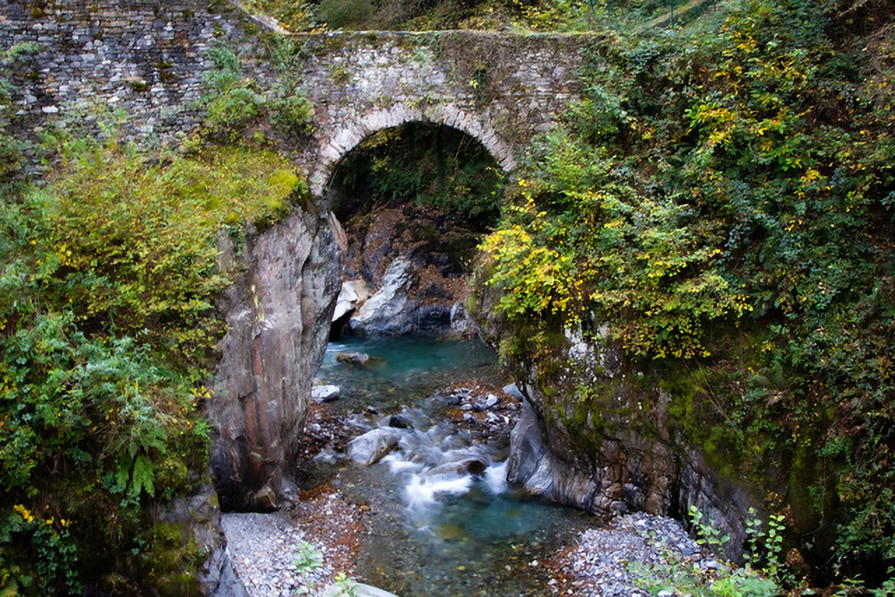 Una pittoresca valle con un ponte di pietra che attraversa un ruscello. Alberi verdi e gialli circondano la zona, mentre l'acqua del ruscello scorre tra le rocce.