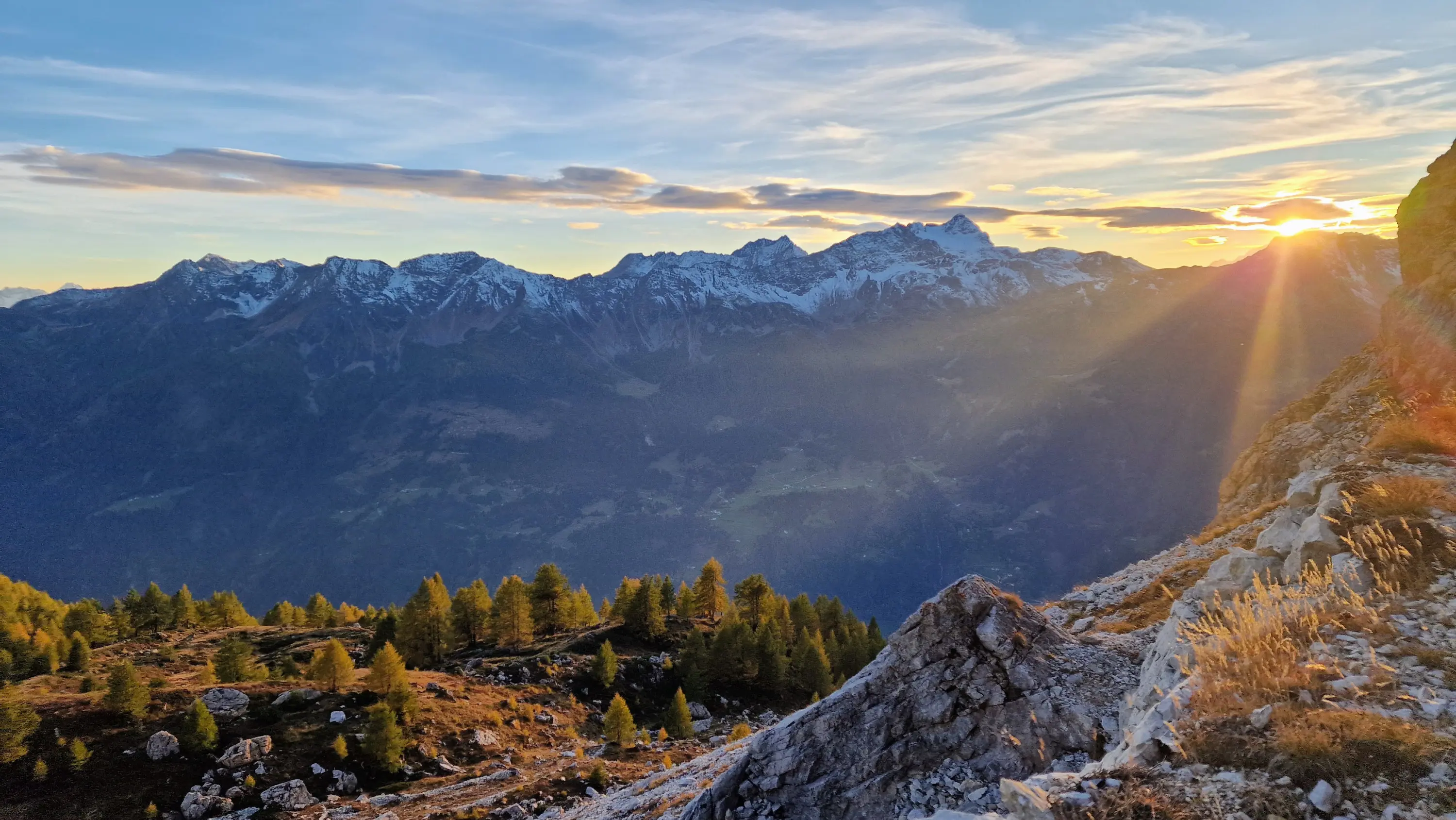 Panorama montano al tramonto, con cime innevate e nuvole nel cielo. La luce del sole si riflette sulle montagne, mentre la vegetazione verdeggiante si estende in primo piano.