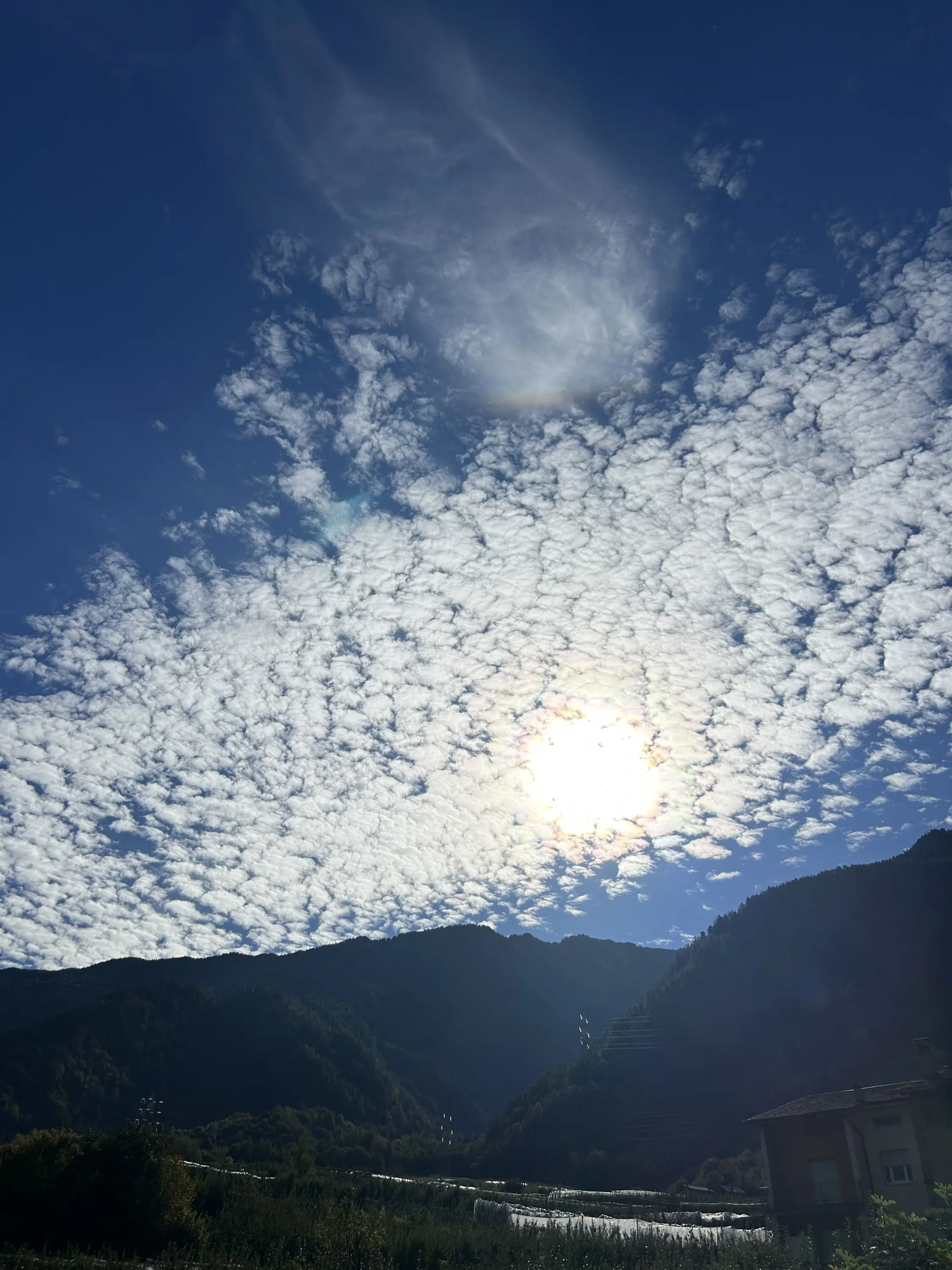 Cielo blu punteggiato da nuvole bianche a strisce, con il sole luminoso che irradia sopra le montagne verdi e una costruzione visibile in basso.