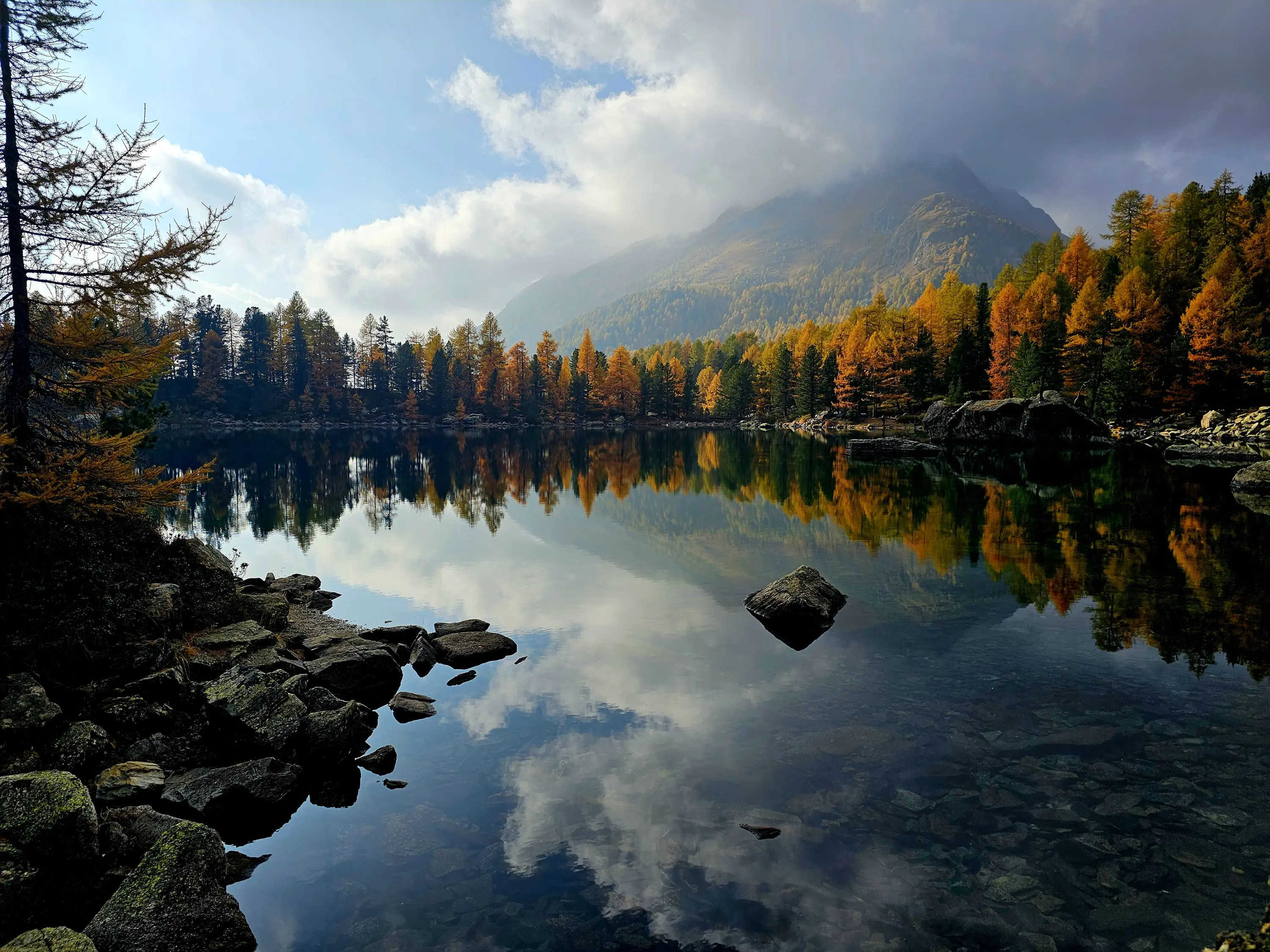 Un lago tranquillo circondato da alberi autunnali. Le montagne si riflettono nell'acqua calmissima, creando un'atmosfera serena. Nuvole e blu del cielo completano il paesaggio.