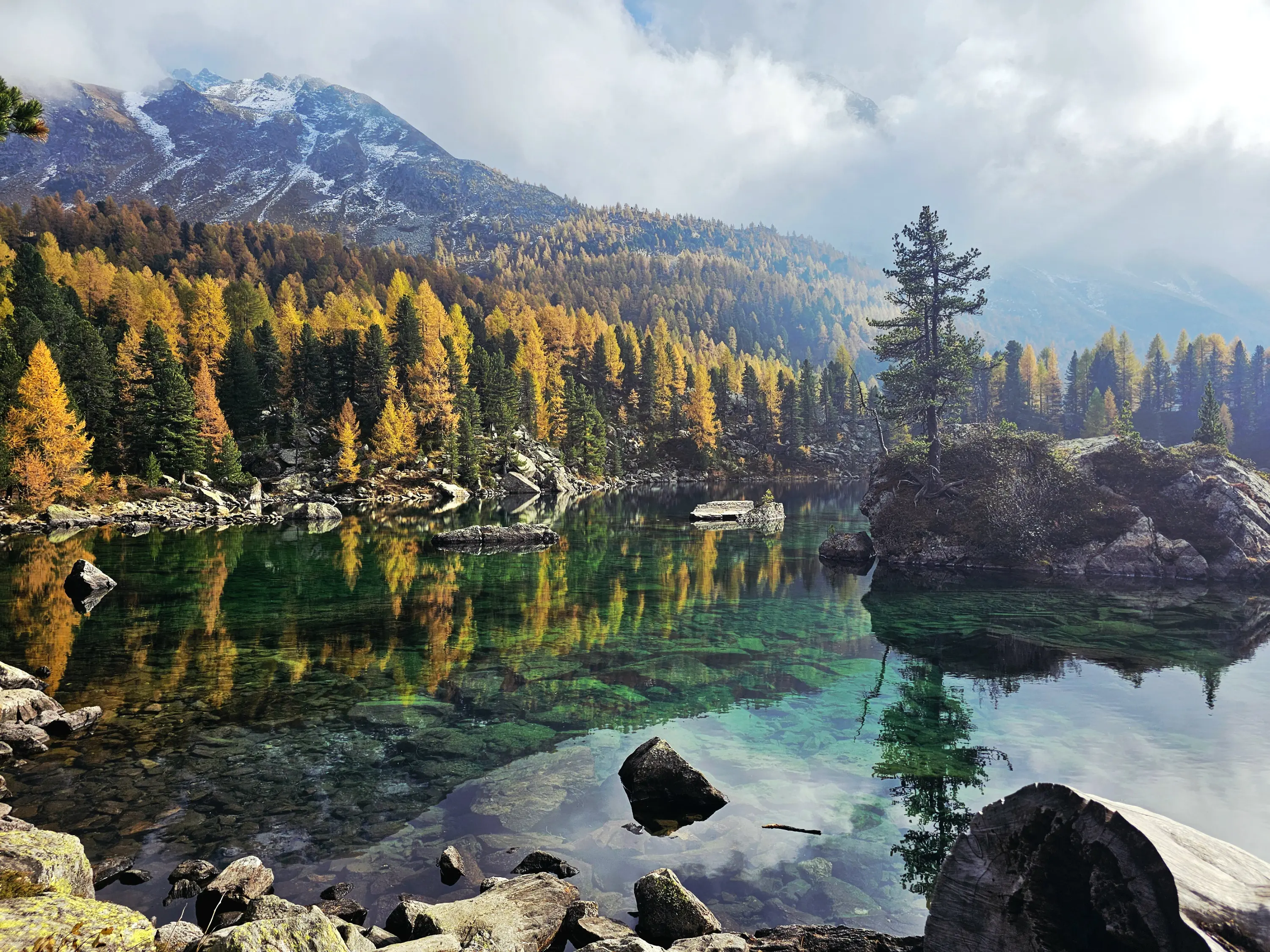 Uno splendido lago circondato da alberi verdi e gialli in autunno, con montagne sullo sfondo e nuvole nel cielo. L'acqua è cristallina e riflette il paesaggio circostante.