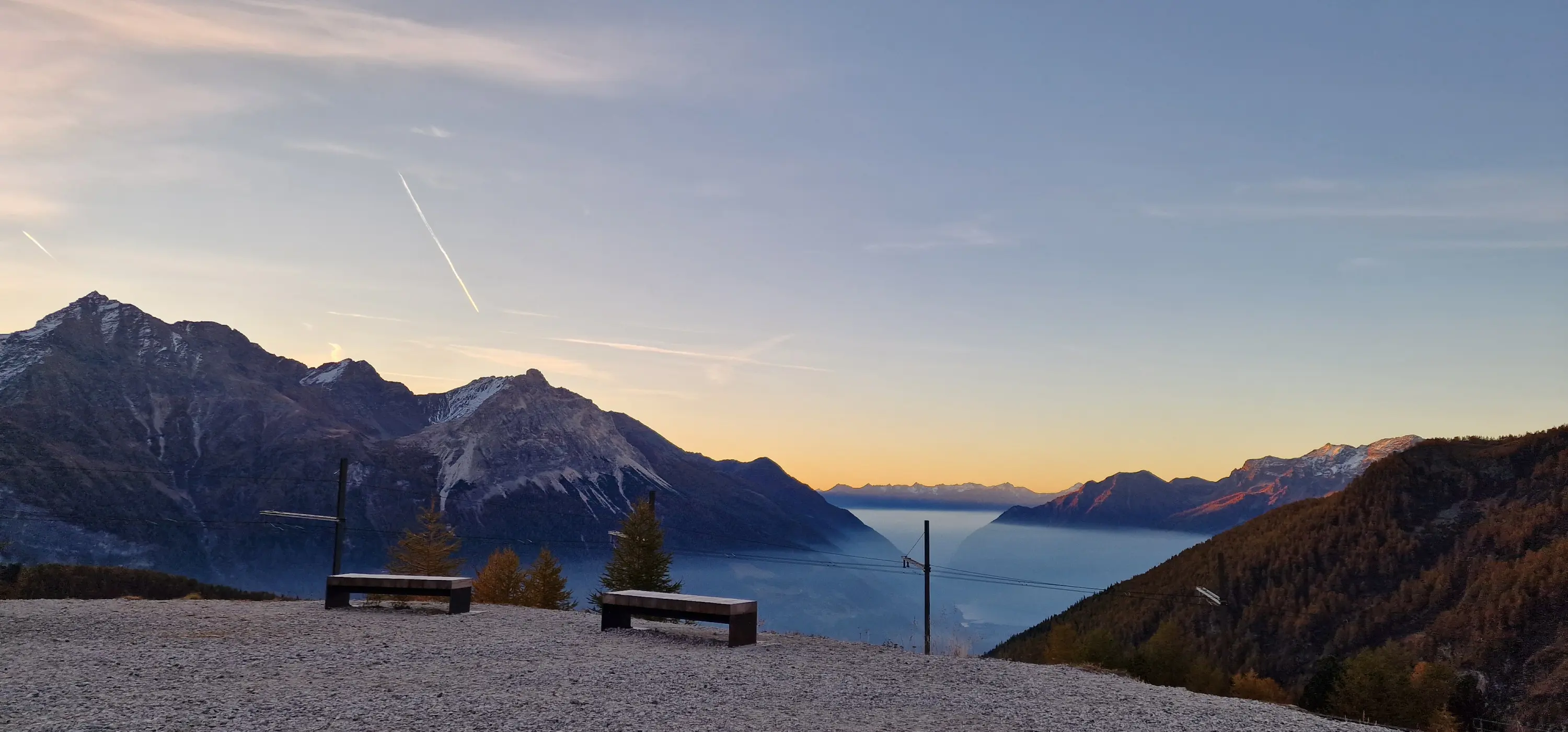 Panorama montano al tramonto, con panchine in legno e valli nebbiose sotto le cime delle montagne. Il cielo è dipinto di tonalità calde.