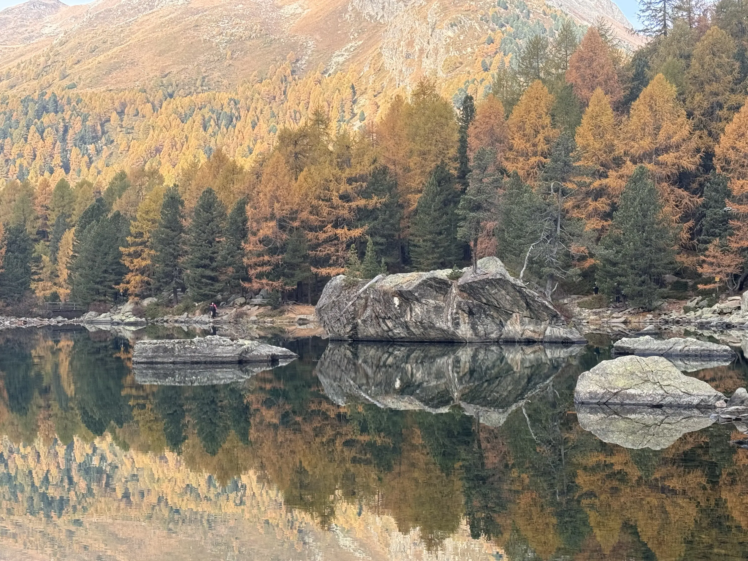 Un paesaggio montano con un lago tranquillo circondato da alberi dai colori autunnali. Le rocce emergono dall'acqua, riflettendo il foliage circostante. Sullo sfondo, le montagne presentano tonalità di marrone e verde.