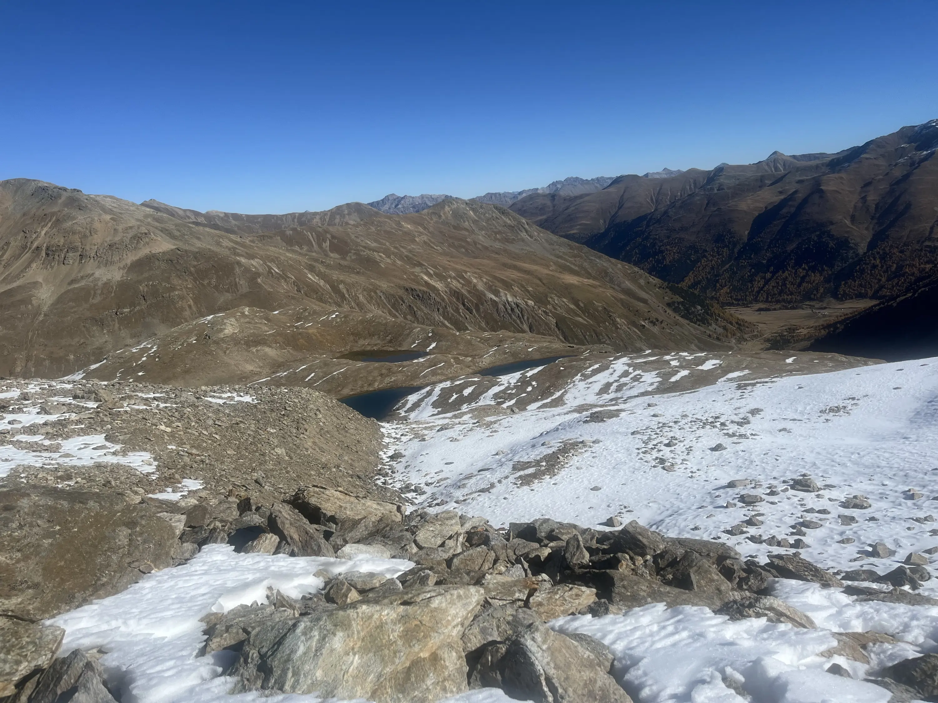 Paesaggio montano con colline brulle e coperte di neve, sotto un cielo blu terso. Sullo sfondo, vette montuose all'orizzonte.