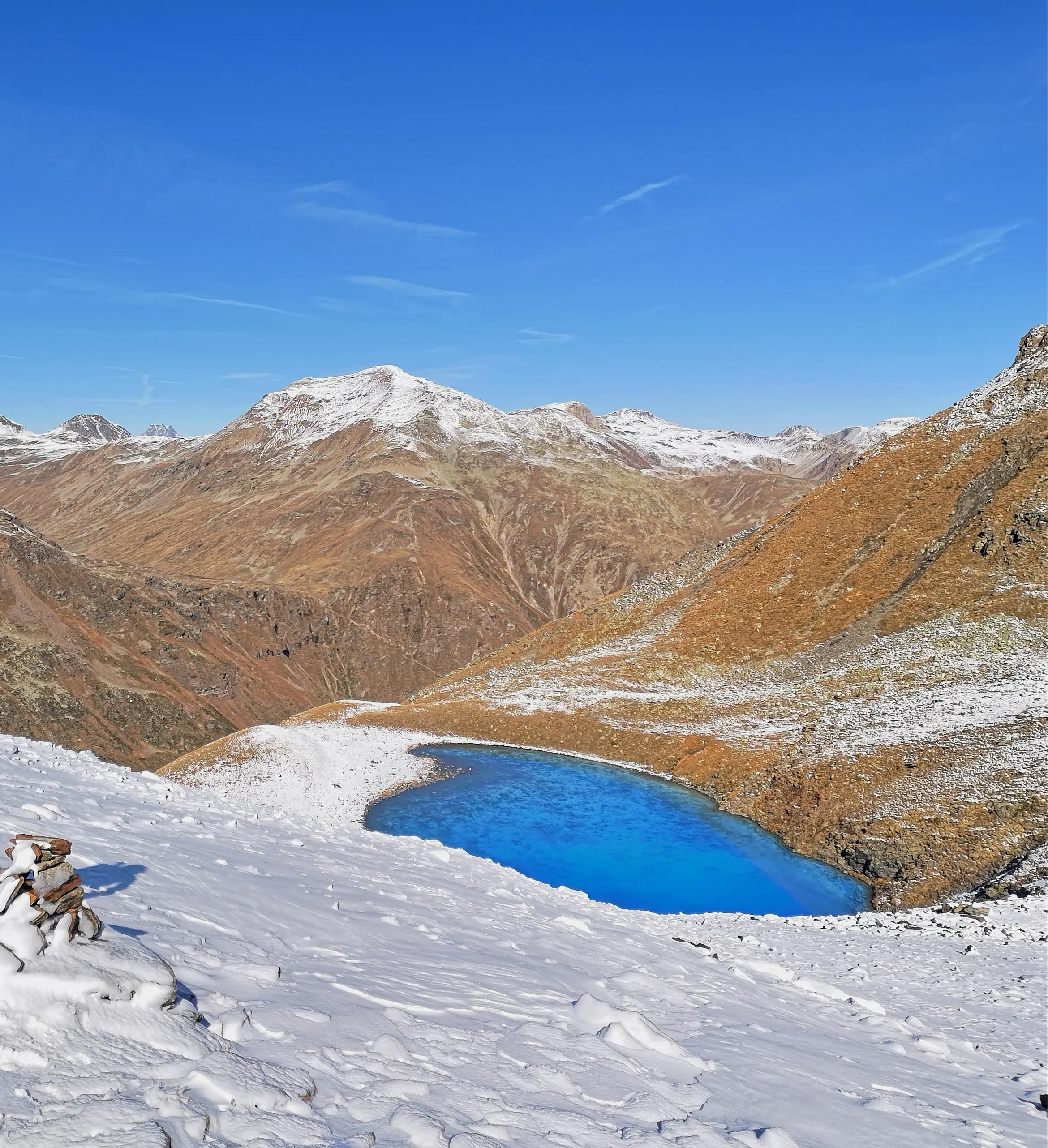 Un lago blu intenso si trova tra monti innevati, circondato da un paesaggio montano dorato e asciutto. Il cielo è sereno e di un blu profondo.