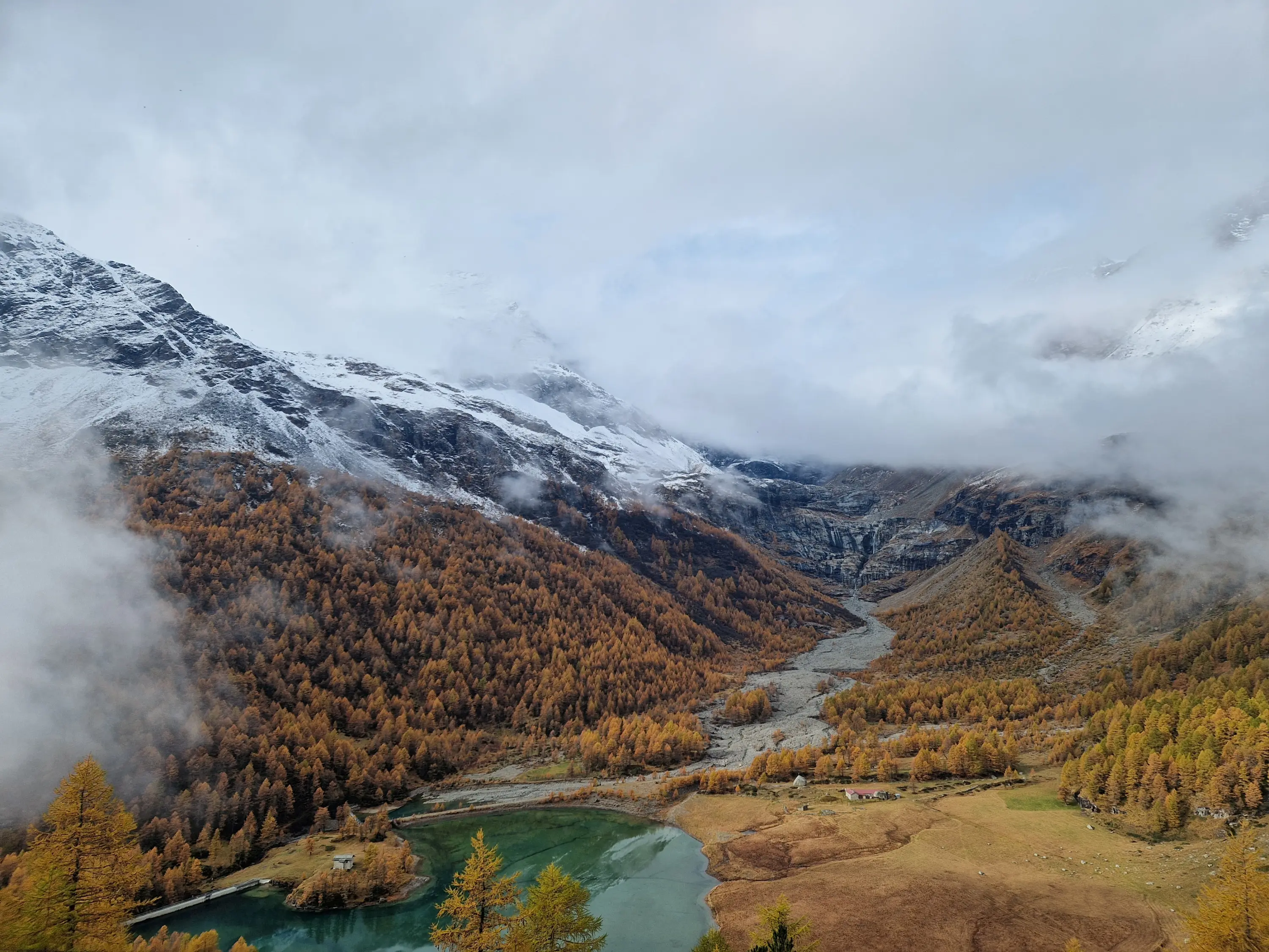 Paesaggio montano autunnale con alberi dorati, una valle che si snoda tra le montagne e un lago verde smeraldo, avvolto da nuvole e nebbia.
