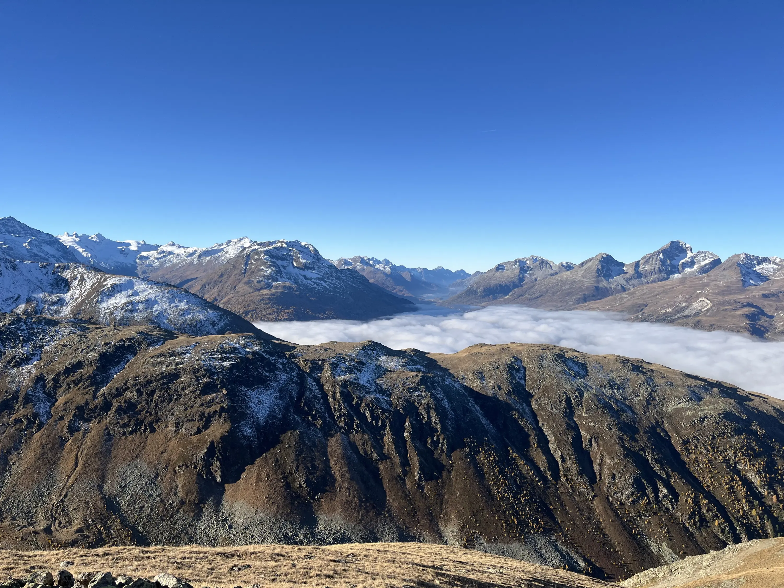 Panorama montano con cime innevate e una vallata coperta di nebbia, sotto un cielo azzurro sereno.