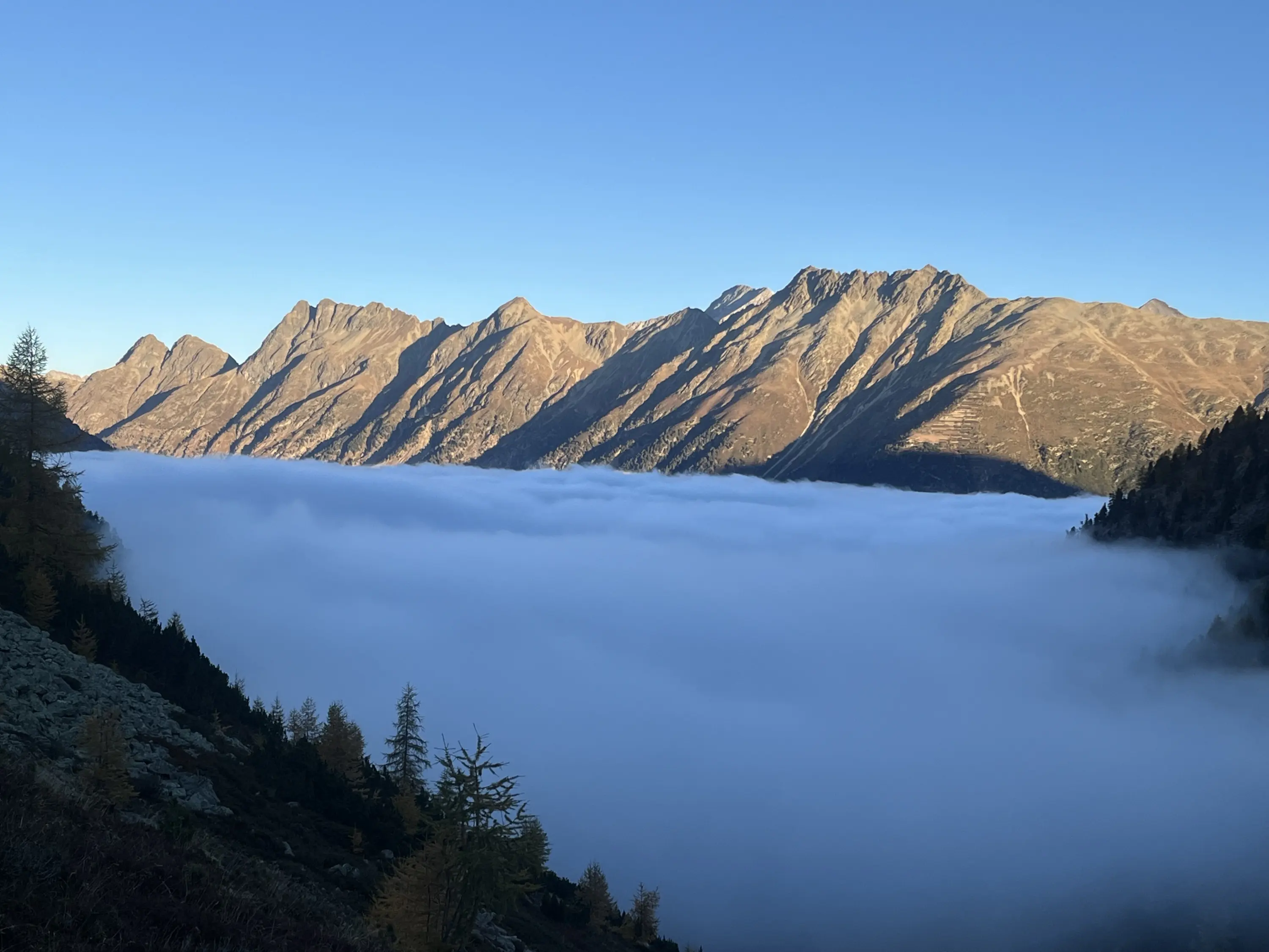 Le montagne si ergono maestose sopra un mare di nuvole dense, creando un contrasto suggestivo con il cielo blu chiaro.