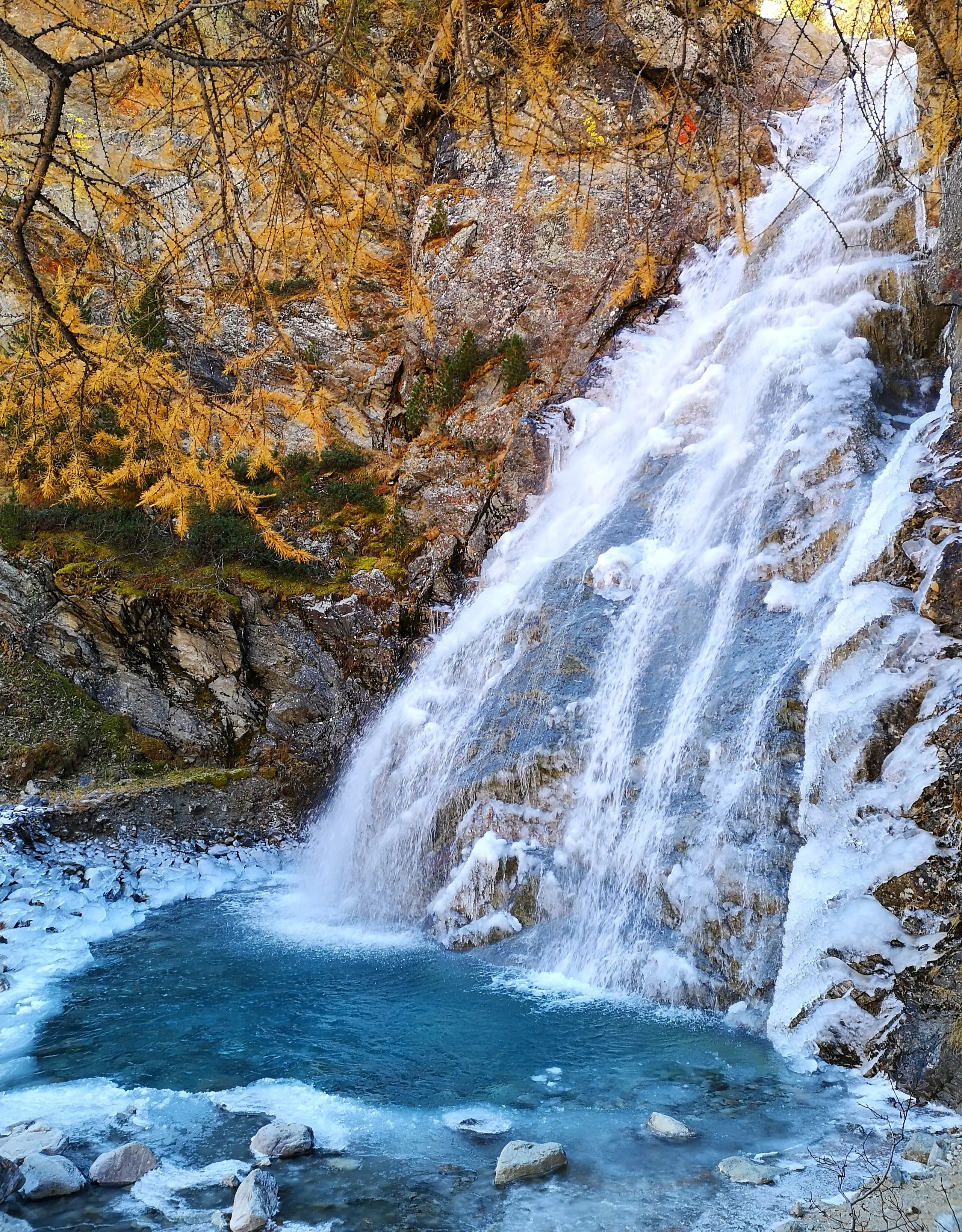 Una cascata che scorre su una roccia, con acqua limpida che si tuffa in una piscina blu sottostante. Attorno, vegetazione e alberi dai colori autunnali.