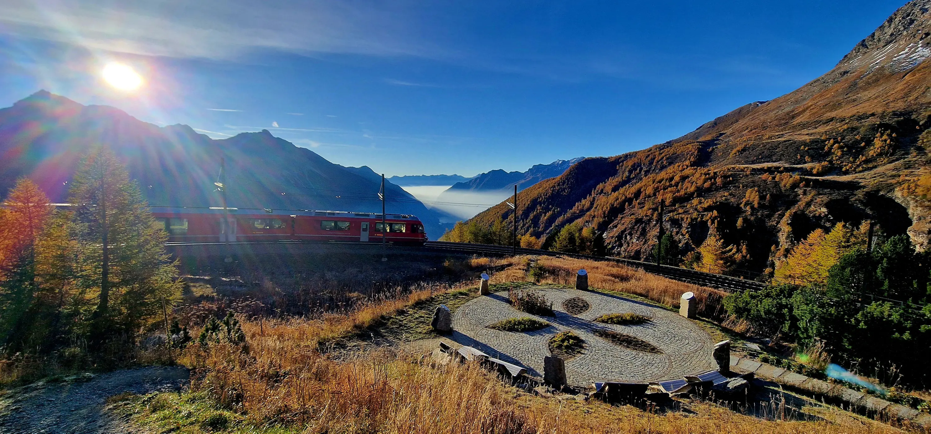 Un treno rosso attraversa un paesaggio montano autunnale, circondato da alberi dorati. Sullo sfondo si vedono le montagne e un lago, con il sole che splende sopra l'orizzonte.
