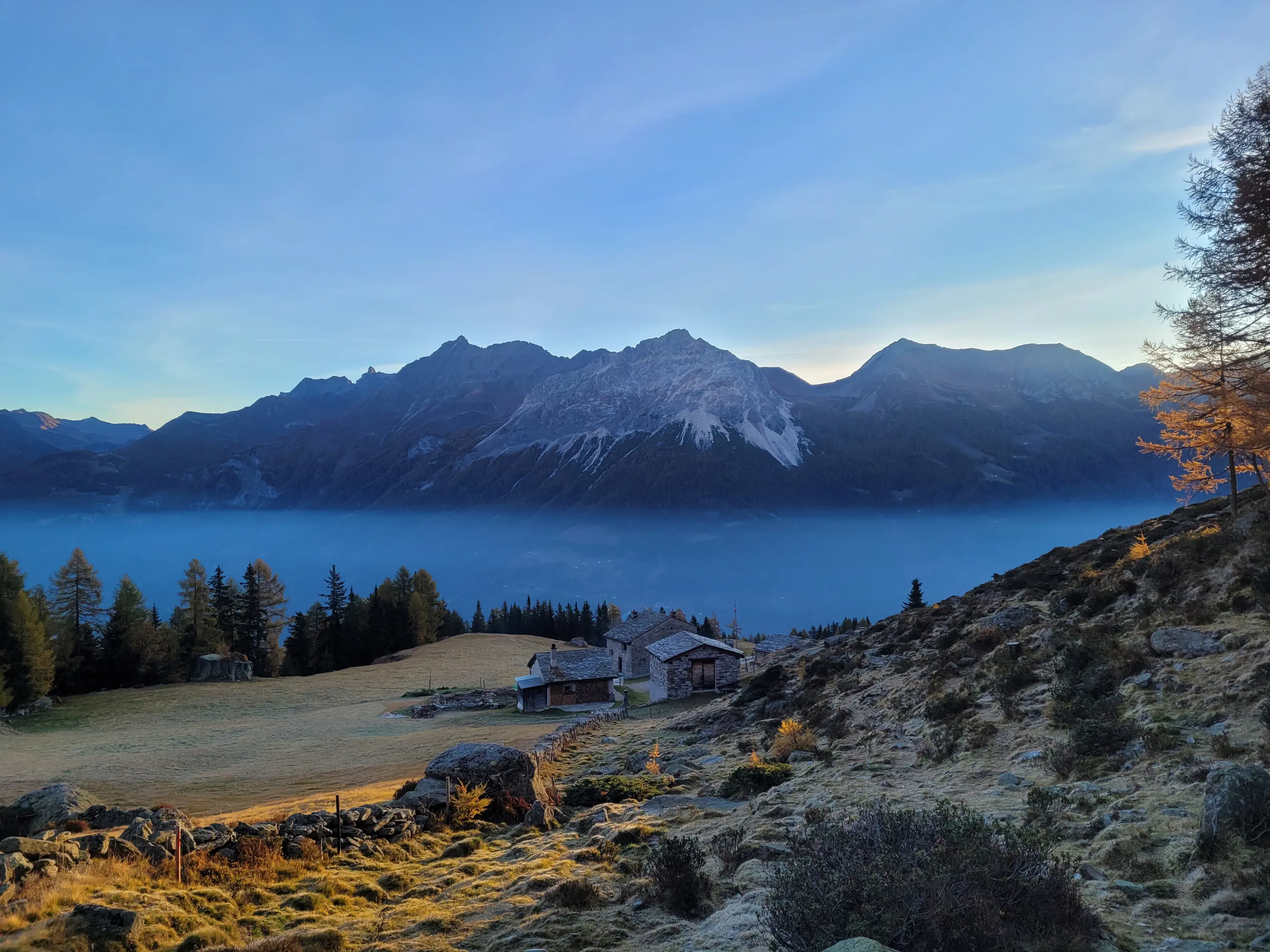 Paesaggio montano al tramonto, con montagne maestose sullo sfondo, deliziose baite sparse su un prato verde e alberi di conifere. La nebbia avvolge la valle sottostante.