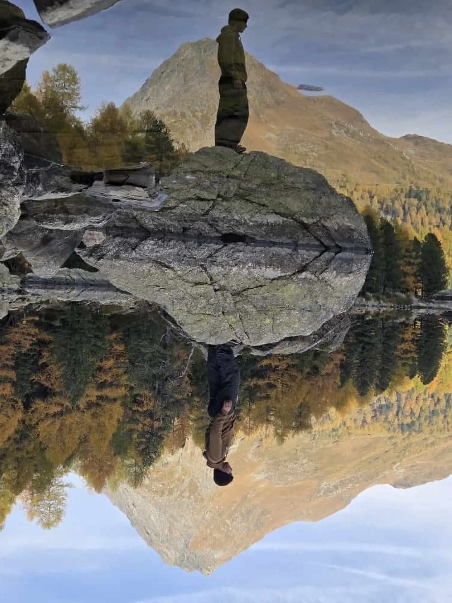 Un uomo in piedi su una grande roccia riflessa in uno stagno circondato da alberi e montagne. Il paesaggio autunnale presenta alberi dai colori dorati e un cielo sereno.