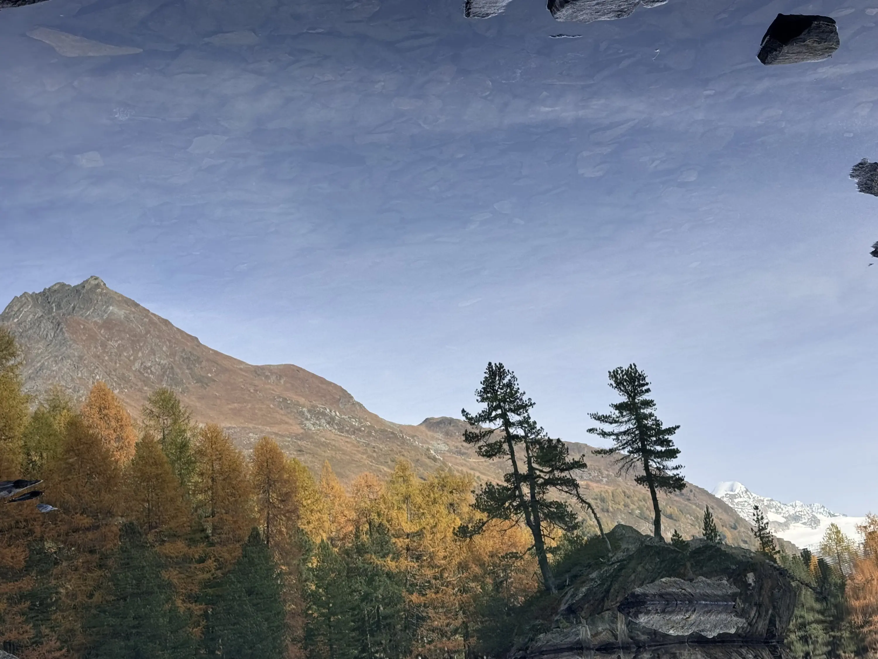 Riflesso di un paesaggio montano con alberi verdi e arancioni in autunno, montagne sullo sfondo e cielo sereno.